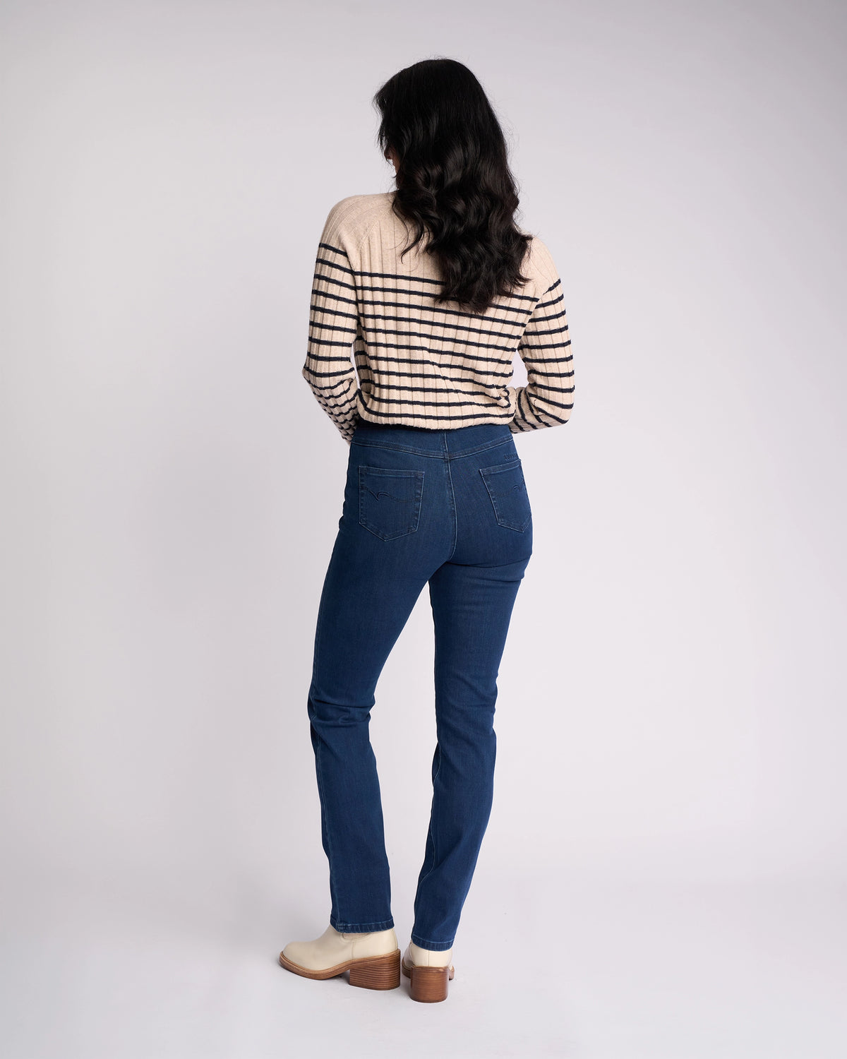 A person with long dark hair, wearing a beige and black striped sweater, Vassalli Bay Pull On - Blue Denim (Without Front Fade) jeans, and light-colored heeled shoes stands facing away from the camera against a plain white background.