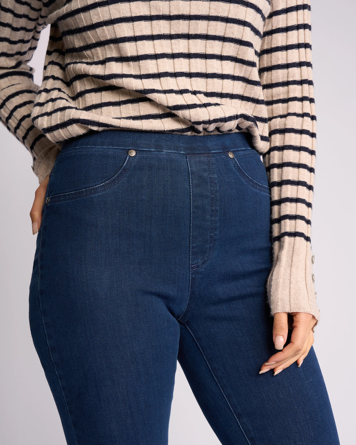 A person wears a beige and black striped sweater with Vassalli Bay Pull On - Blue Denim (Without Front Fade) jeans, shown from shoulders to mid-thigh, with one hand resting by their side.