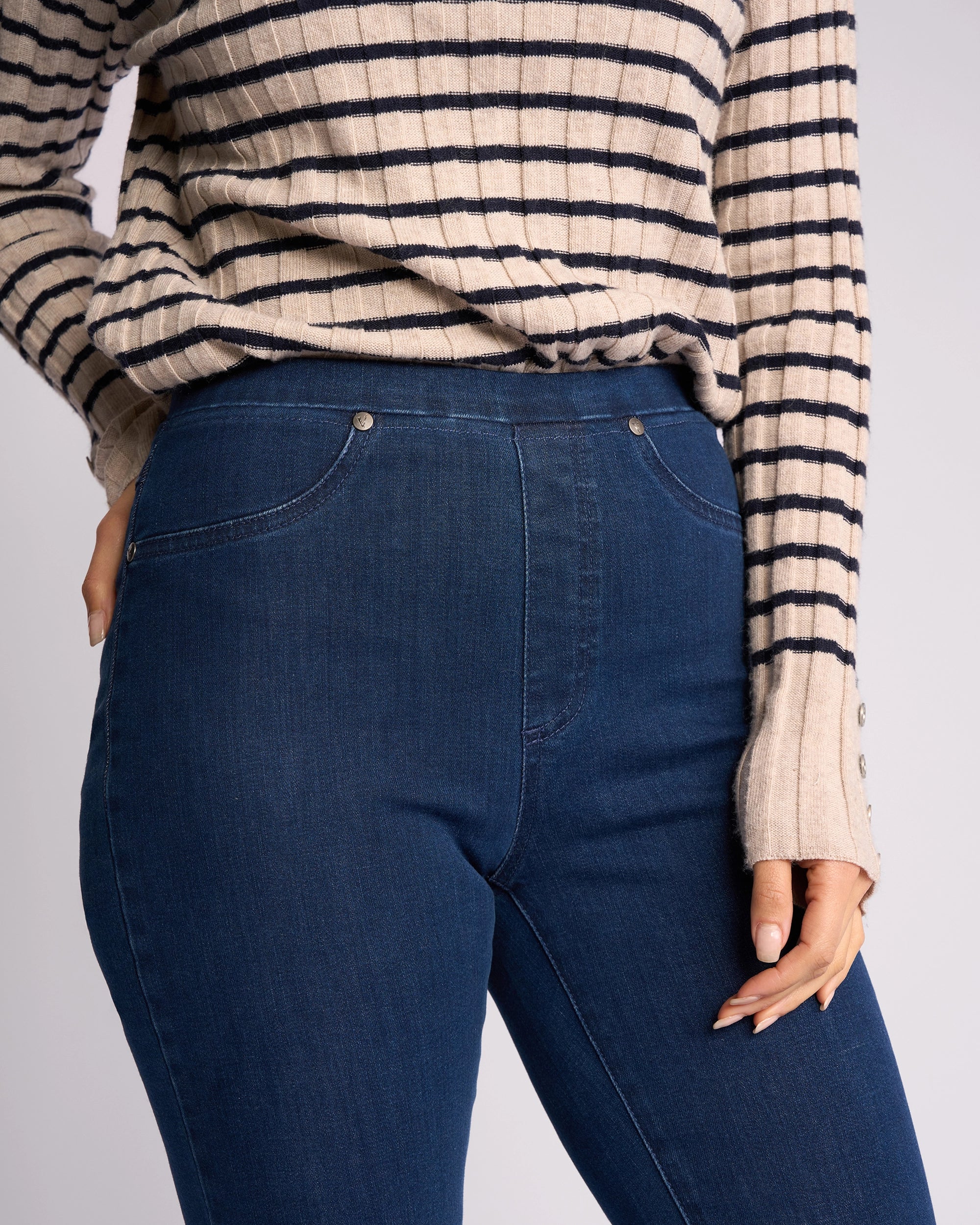 A person wears a beige and black striped sweater with Vassalli Bay Pull On - Blue Denim (Without Front Fade) jeans, shown from shoulders to mid-thigh, with one hand resting by their side.