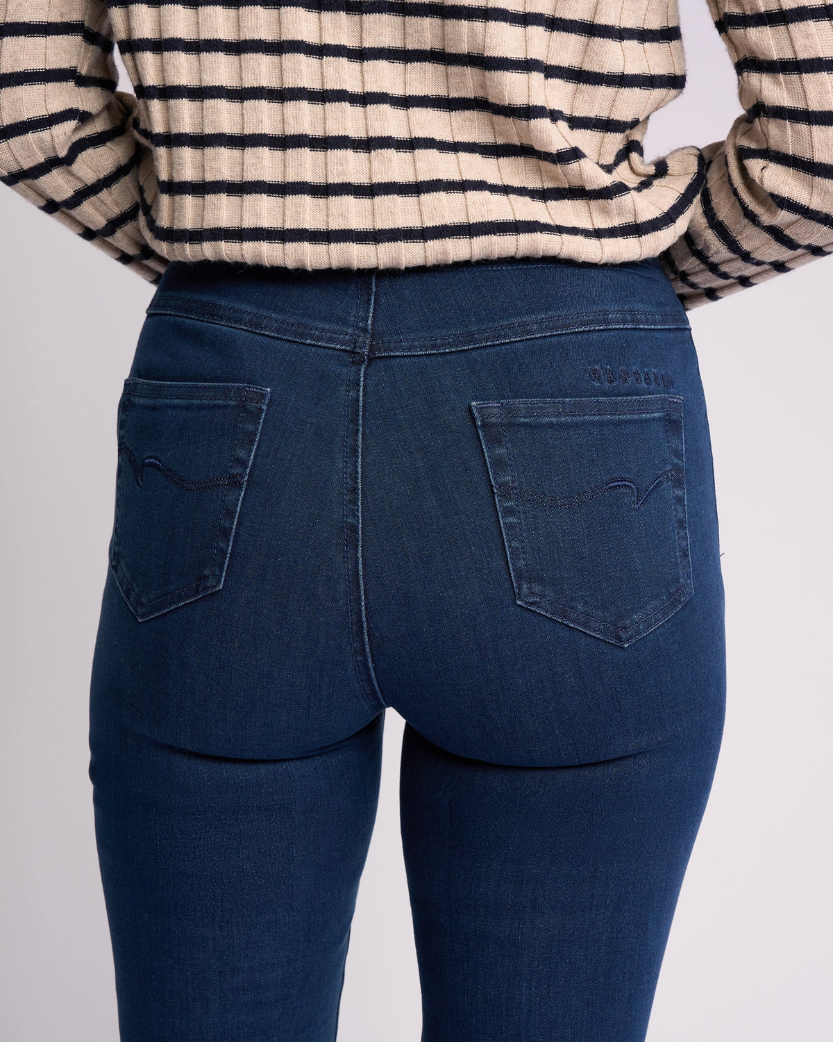 A person is shown from the back wearing Vassalli's Bay Pull On - Blue Denim (Without Front Fade) jeans, highlighting the slim-leg pockets and lower back, paired with a beige long-sleeve, black-striped top.