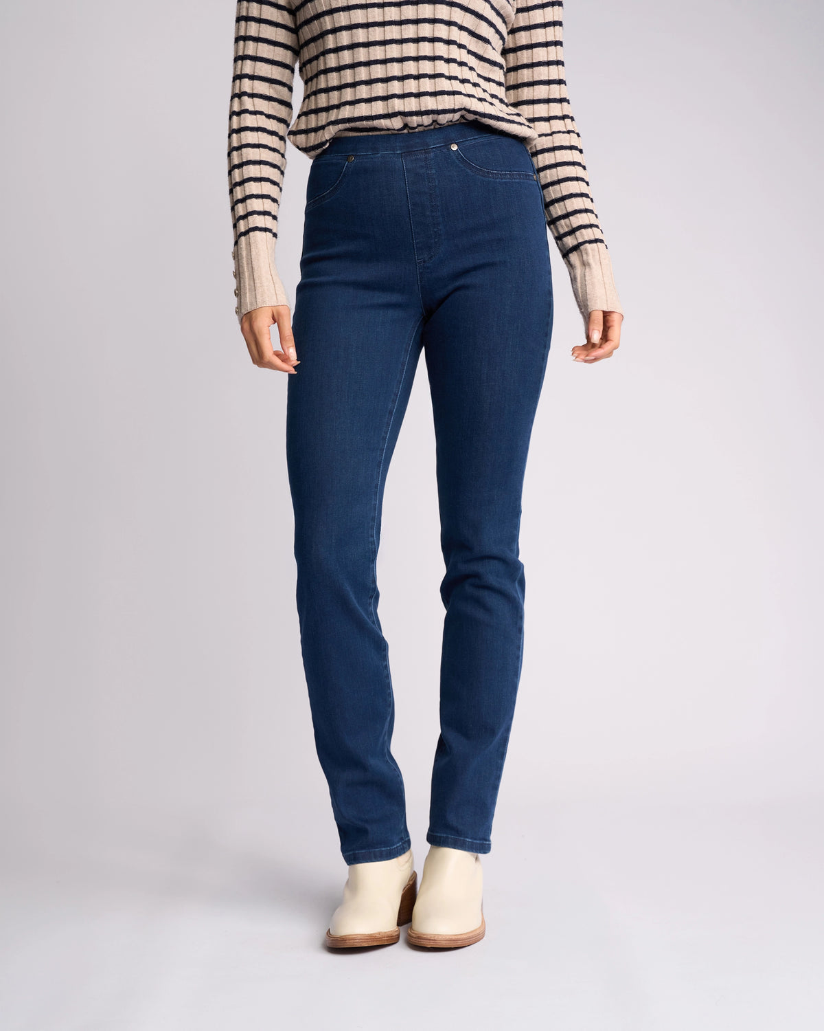 A person stands against a neutral background wearing Vassalli's Bay Pull On - Blue Denim (Without Front Fade) jeans. They pair the high-rise slim-leg jeans with a striped long-sleeve shirt and cream ankle boots; their head is not visible.