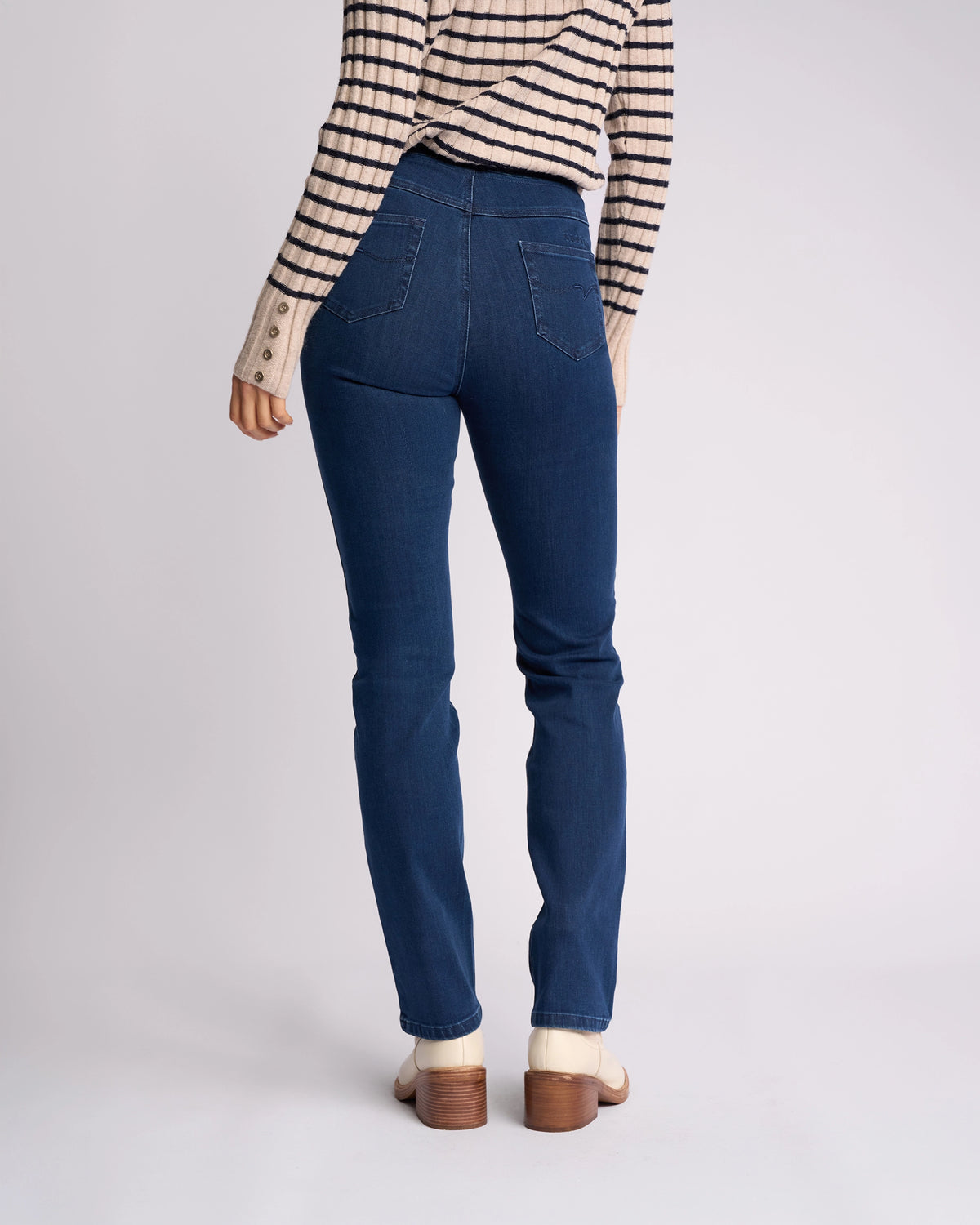 A person stands facing away, wearing Vassalli's Bay Pull On - Blue Denim (Without Front Fade) jeans, a striped long-sleeve shirt, and cream boots, showcasing the back view of the outfit against a plain light background.