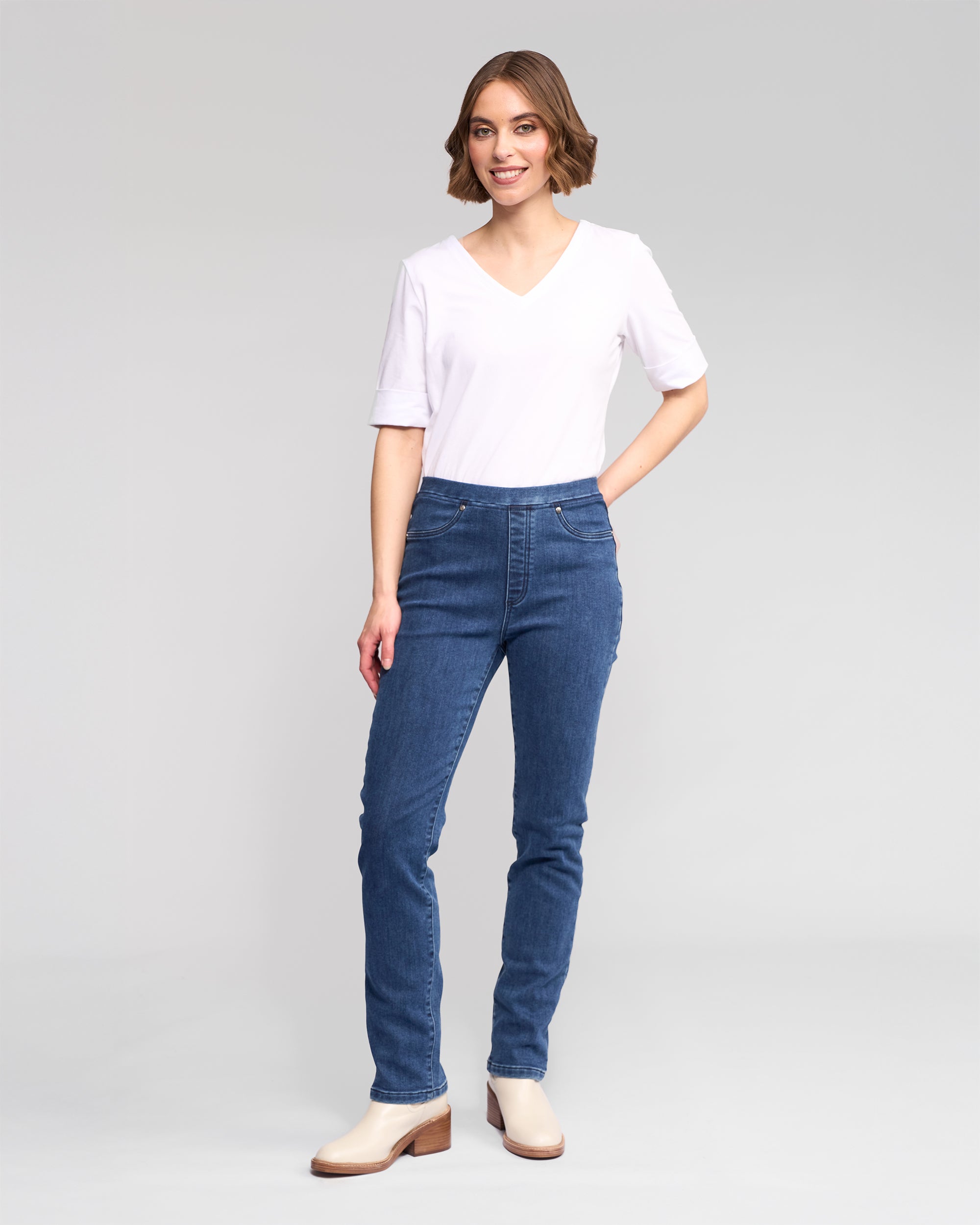 A woman with short brown hair smiles, wearing a white V-neck tee and Vassalli's Bay Pull On - Brushed Indigo denim pants, paired with beige ankle boots. She poses against a plain light gray background.
