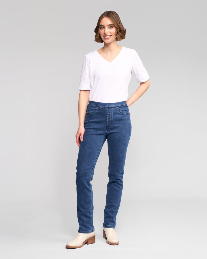 A woman with short brown hair smiles, wearing a white V-neck tee and Vassalli's Bay Pull On - Brushed Indigo denim pants, paired with beige ankle boots. She poses against a plain light gray background.