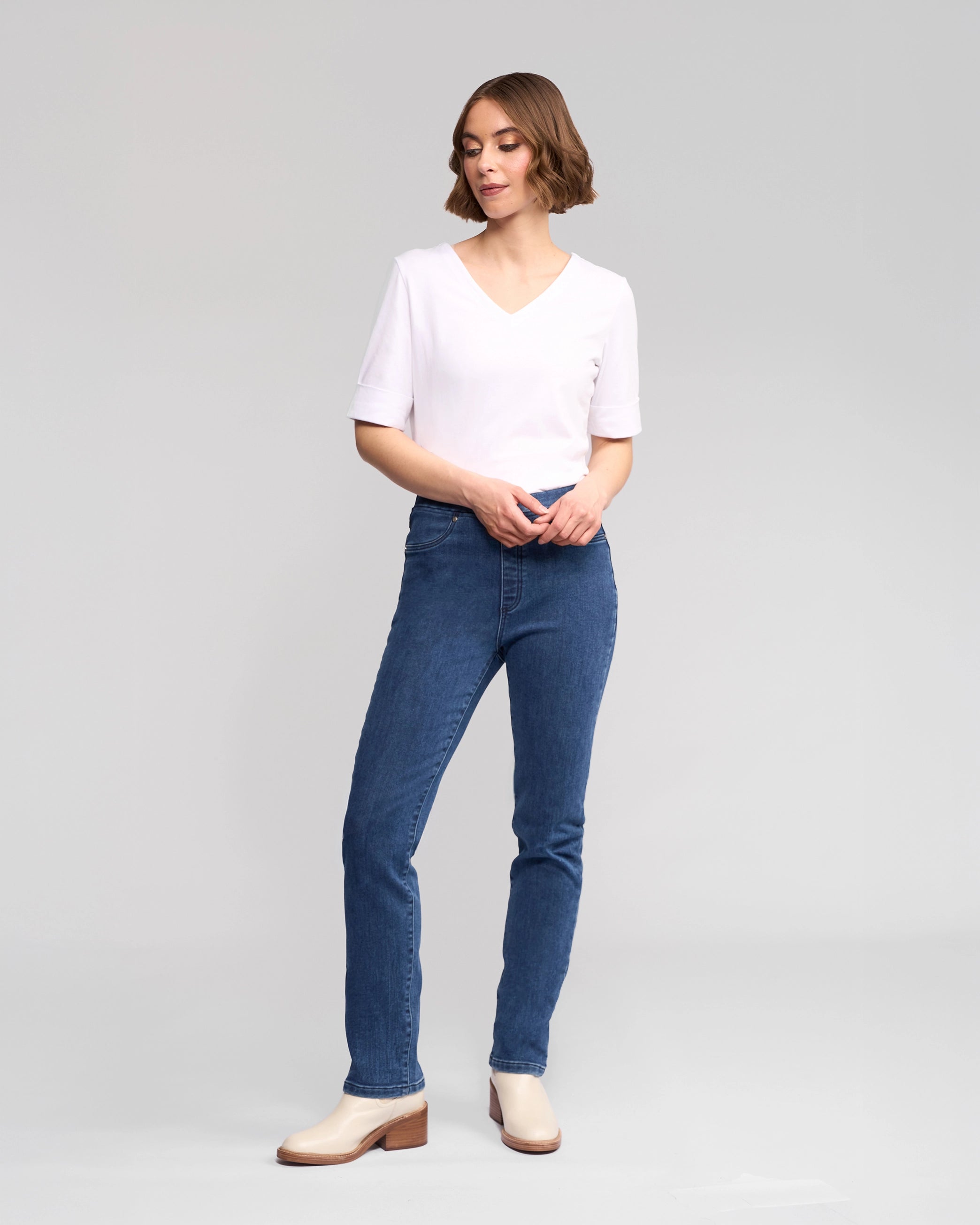 A person stands against a plain background wearing Vassalli's Bay Pull On - Brushed Indigo jeans, a white short-sleeve V-neck shirt, and beige ankle boots, looking slightly to the side with hands loosely clasped in front.