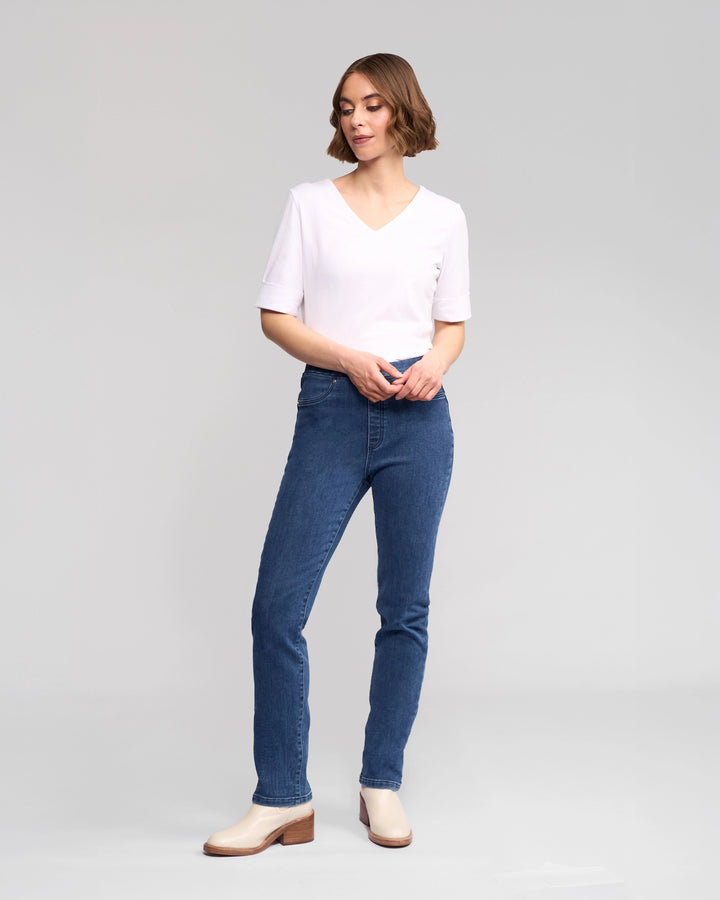 A person stands against a plain background wearing Vassalli's Bay Pull On - Brushed Indigo jeans, a white short-sleeve V-neck shirt, and beige ankle boots, looking slightly to the side with hands loosely clasped in front.
