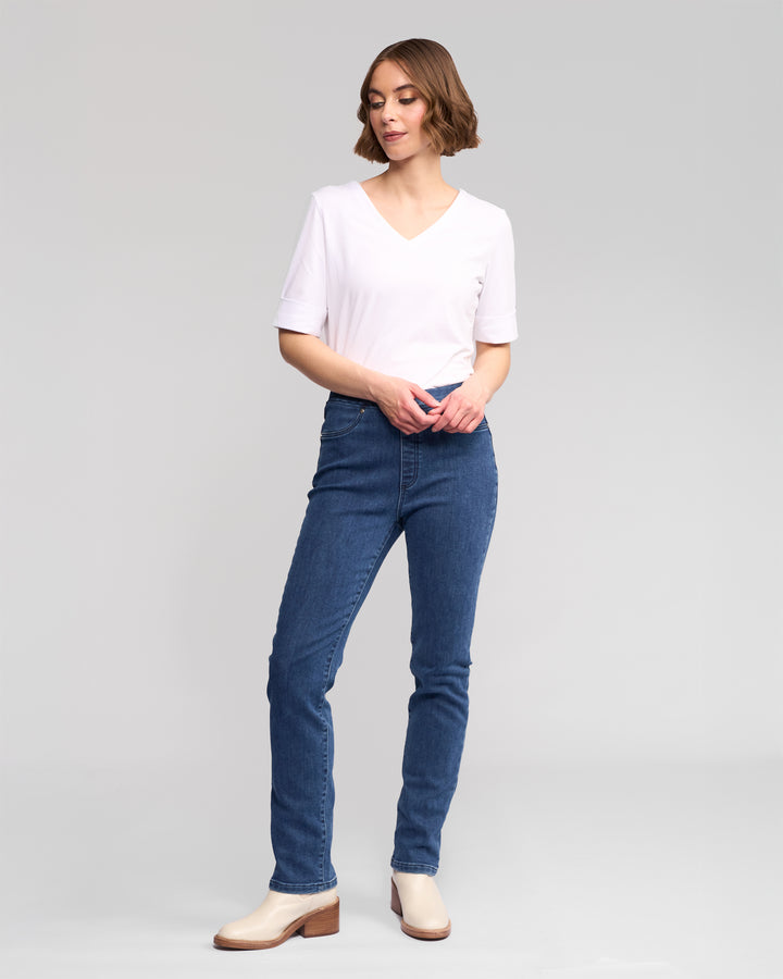 A woman stands against a gray background, wearing Vassalli's Bay Pull On - Brushed Indigo slim stretch denim pants with an elastic waistband, a white V-neck shirt, and light heeled shoes. She gazes to the side, one hand holding her wrist.