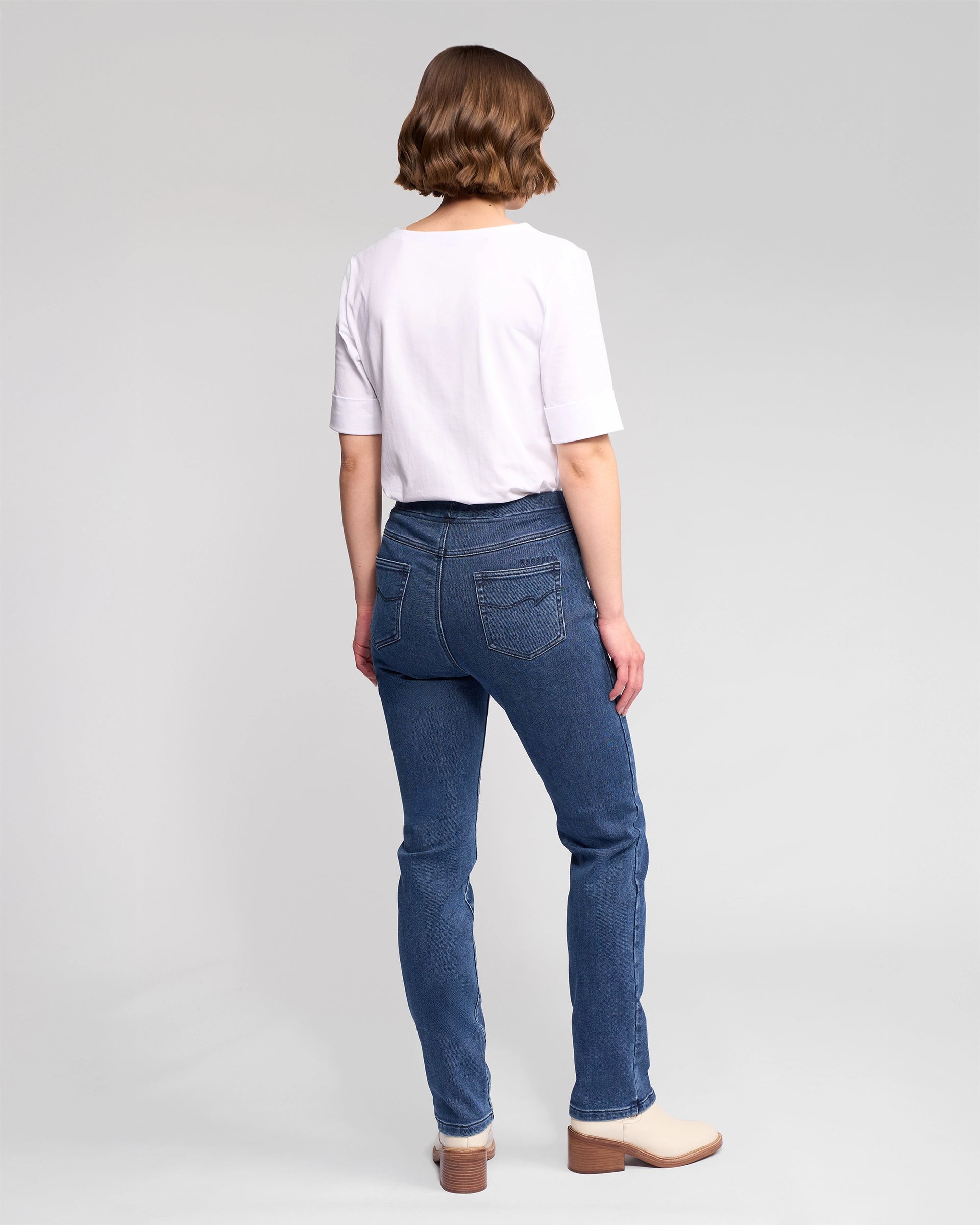 A woman with short brown hair, seen from behind, wears a white short-sleeve top and Vassalli Bay Pull On - Brushed Indigo jeans with beige ankle boots, set against a plain light gray background.