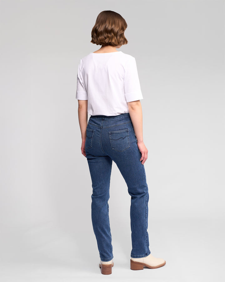 A person with short brown hair stands facing away in a white t-shirt and Vassalli Bay Pull On - Brushed Indigo slim stretch denim pants with an elastic waistband, plus beige ankle boots, against a plain light gray background.