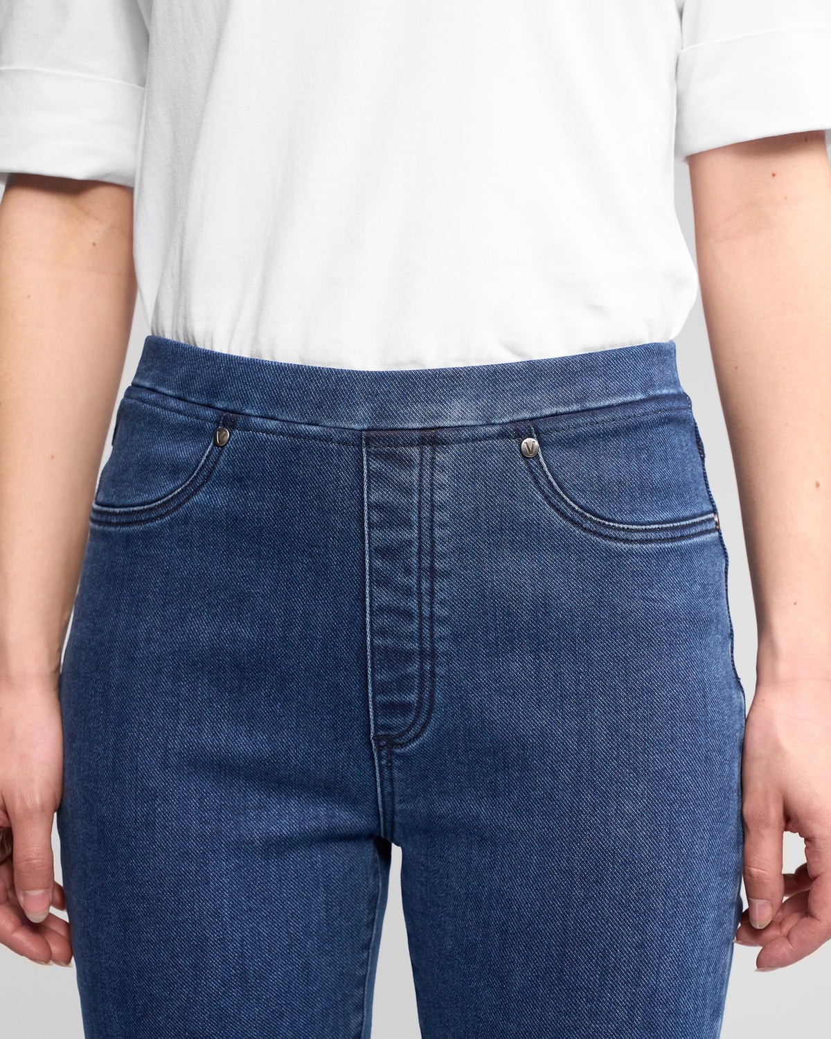 A person wearing a white short-sleeve shirt and Vassalli Bay Pull On - Brushed Indigo high-rise slim-leg jeans, shown from shoulders to mid-thigh with hands relaxed at their sides.