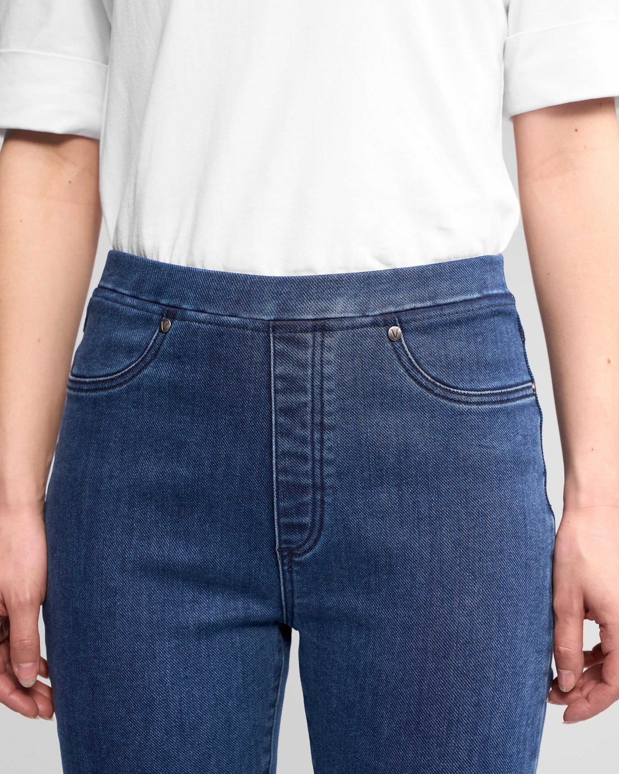 A person wearing a white short-sleeve shirt and Vassalli Bay Pull On - Brushed Indigo high-rise slim-leg jeans, shown from shoulders to mid-thigh with hands relaxed at their sides.