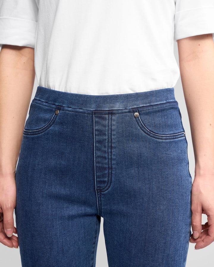 A person wearing a white short-sleeve shirt and Vassalli Bay Pull On - Brushed Indigo high-rise slim-leg jeans, shown from shoulders to mid-thigh with hands relaxed at their sides.