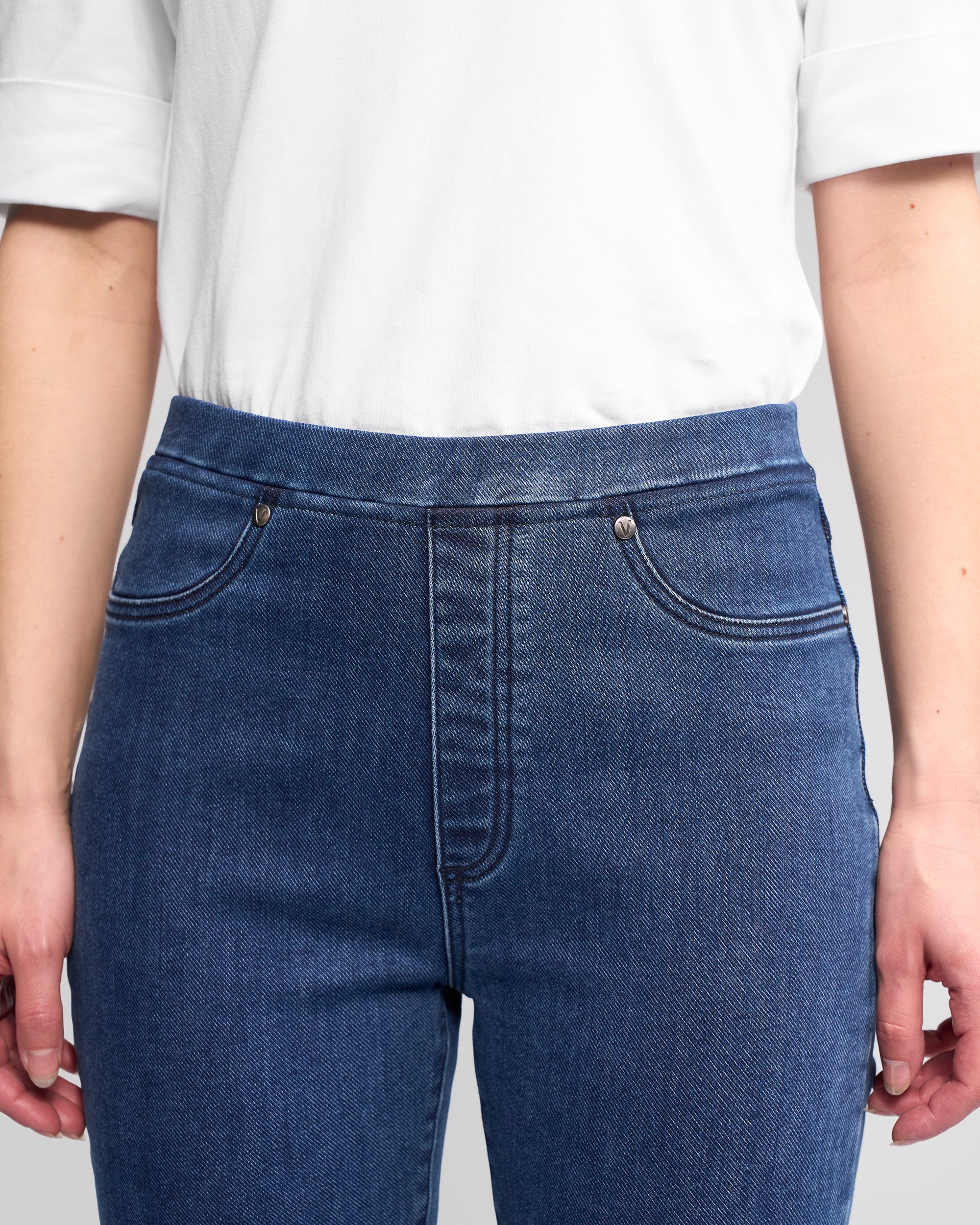 A person wears Vassalli Bay Pull On - Brushed Indigo slim stretch denim pants with an elastic waistband and a white short-sleeve shirt; only their torso and upper legs are visible, with hands relaxed at their sides.