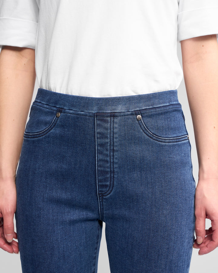 A person wears Vassalli Bay Pull On - Brushed Indigo slim stretch denim pants with an elastic waistband and a white short-sleeve shirt; only their torso and upper legs are visible, with hands relaxed at their sides.