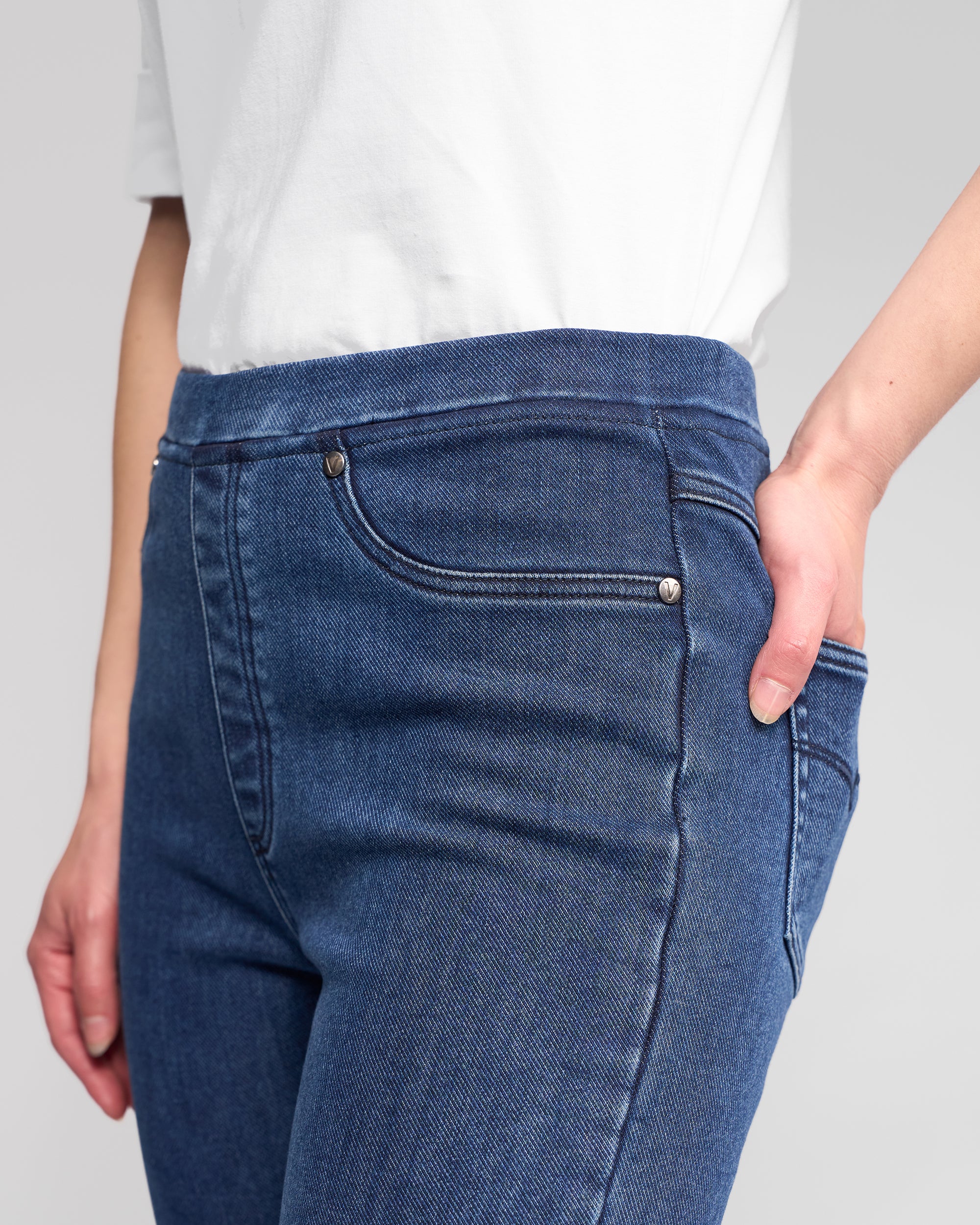 Someone wearing Vassalli Bay Pull On pants in Brushed Indigo, featuring a slim stretch fit and elastic waistband, is shown from the waist down with one hand in their pocket.