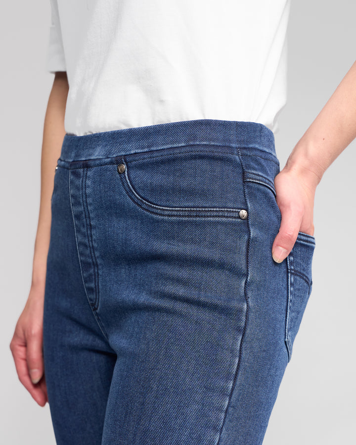 Someone wearing Vassalli Bay Pull On pants in Brushed Indigo, featuring a slim stretch fit and elastic waistband, is shown from the waist down with one hand in their pocket.
