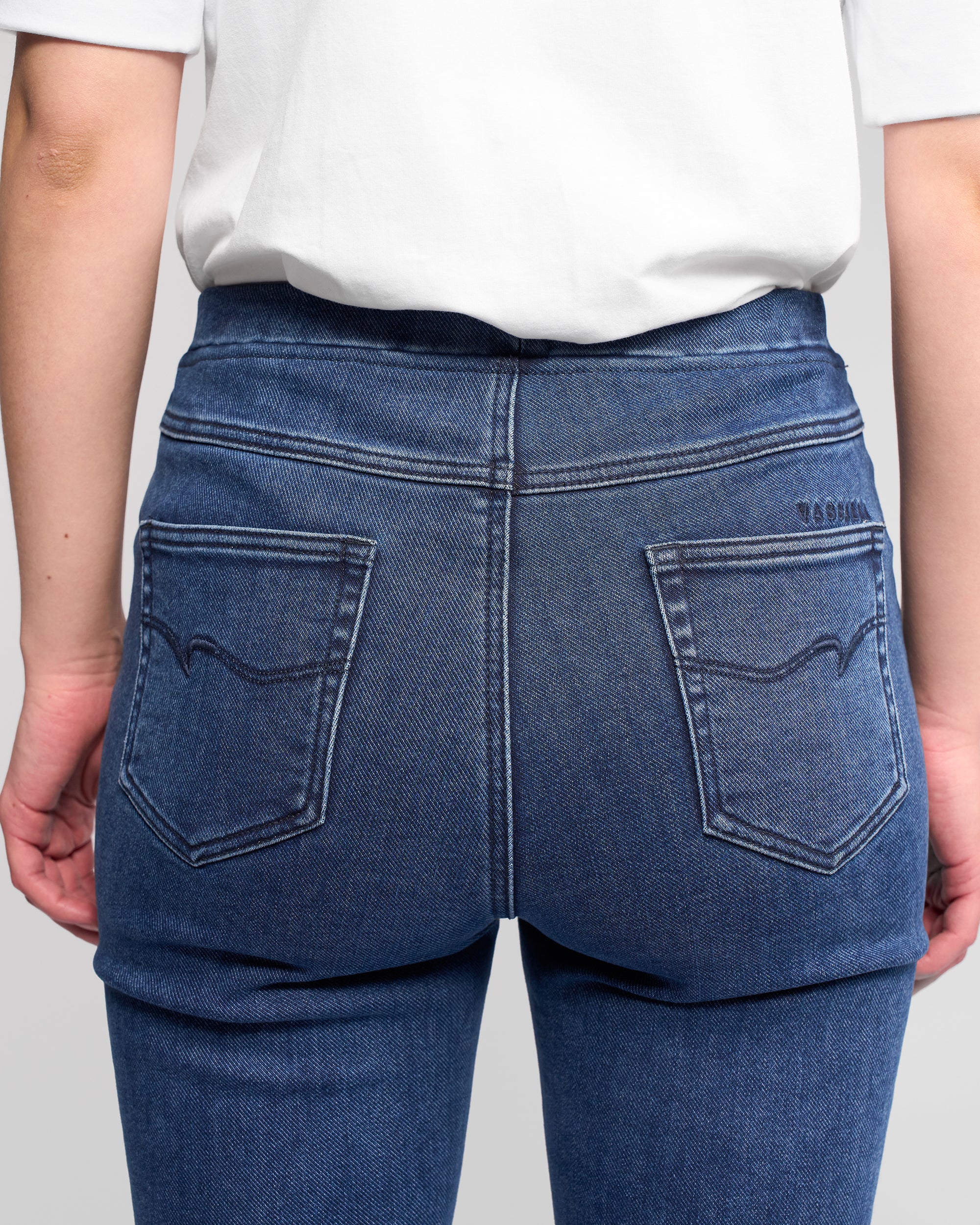 Model wearing Vassalli Bay Pull On - Brushed Indigo, shown from the back with focus on the elastic waistband and back pockets. White shirt and slim stretch fit are highlighted against a plain background.