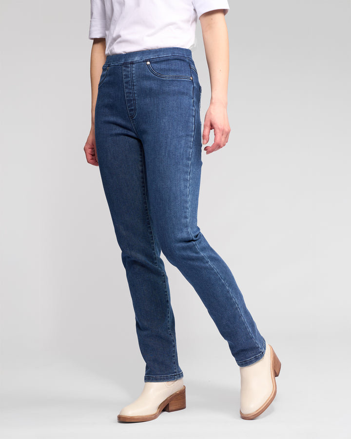 A person stands against a plain light gray background wearing Vassalli's Bay Pull On - Brushed Indigo slim stretch denim pants with an elastic waistband, a white top, and light beige heeled ankle boots. The person's head is not visible.