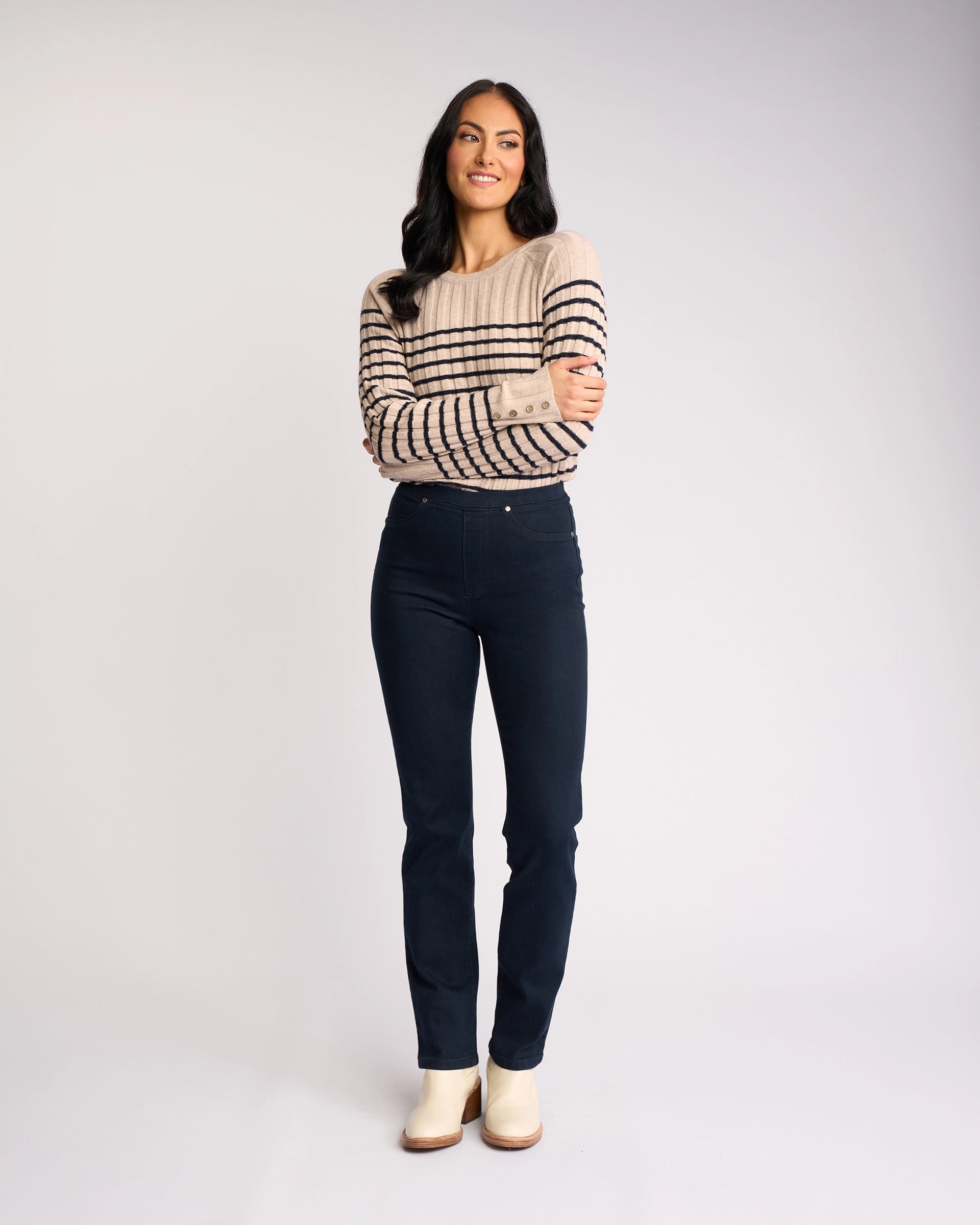 A woman with long dark hair stands, arms crossed and smiling, wearing a beige sweater with black stripes and Vassalli Bay Pull On - Indigo Denim jeans against a plain white background. She looks slightly to the side, finished with light-colored shoes.