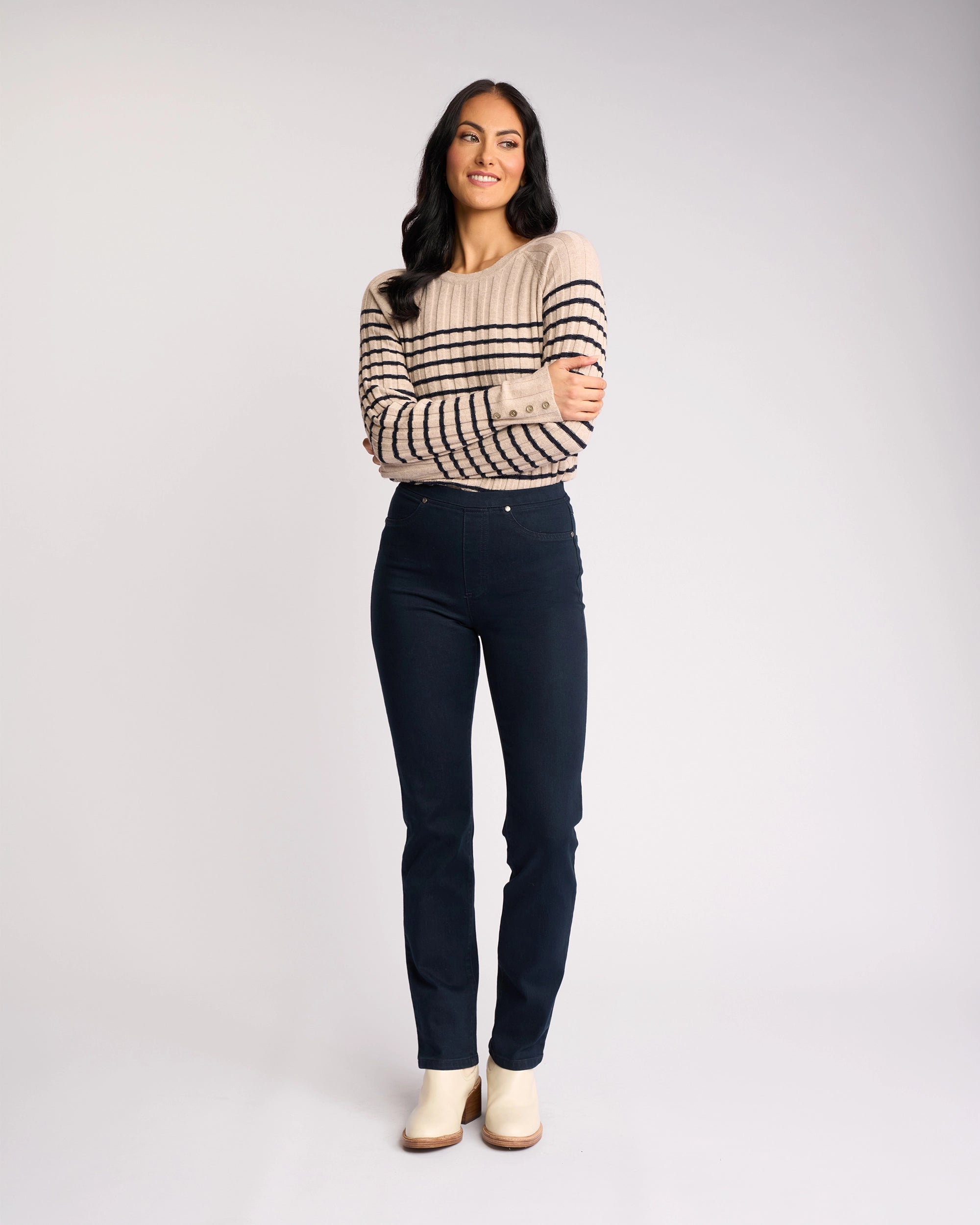 A woman with long dark hair stands, arms crossed and smiling, wearing a beige sweater with black stripes and Vassalli Bay Pull On - Indigo Denim jeans against a plain white background. She looks slightly to the side, finished with light-colored shoes.
