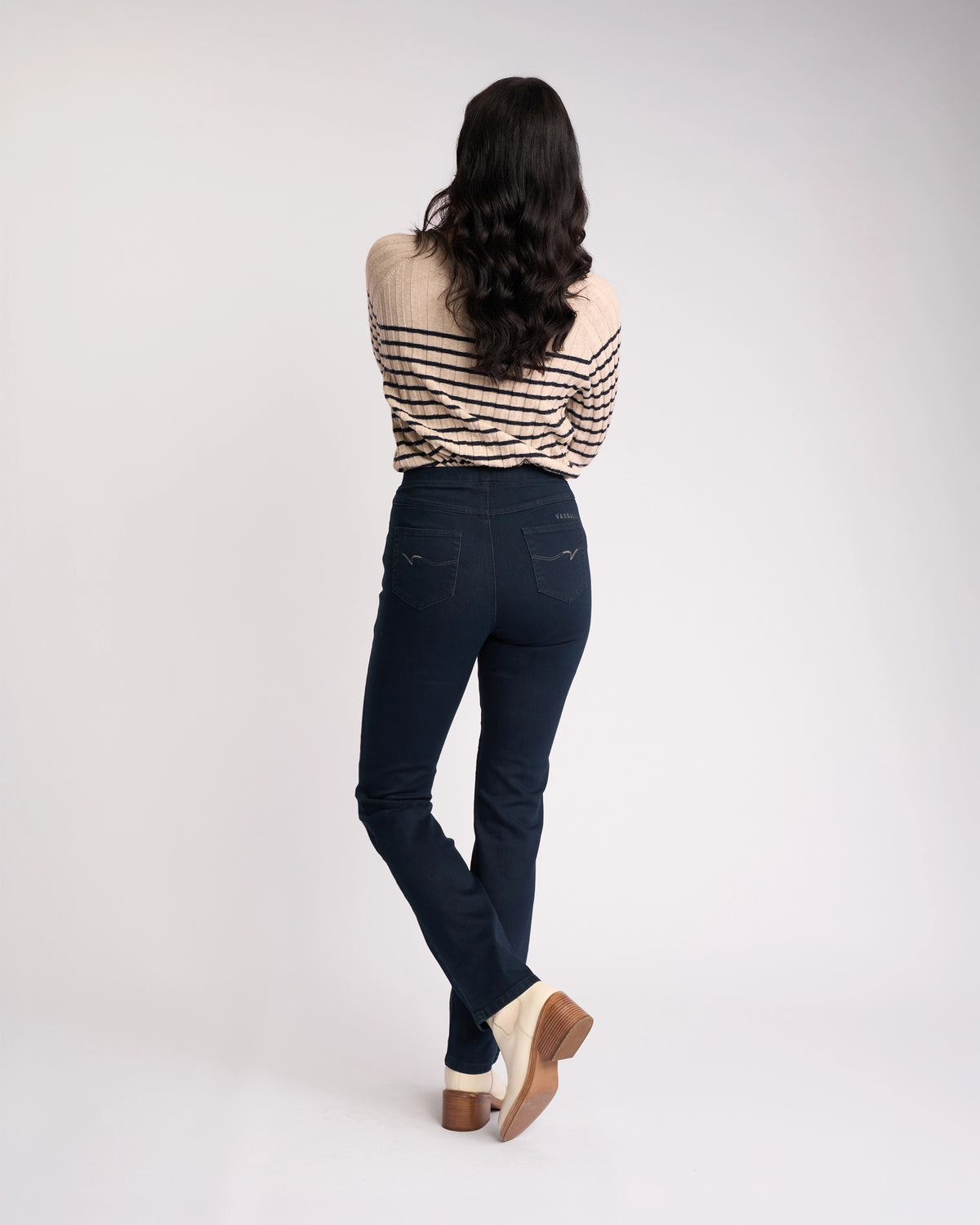 A person with long dark wavy hair stands facing away, wearing a beige sweater with black stripes, Vassalli Bay Pull On - Indigo Denim jeans, and beige heeled shoes against a plain white background.