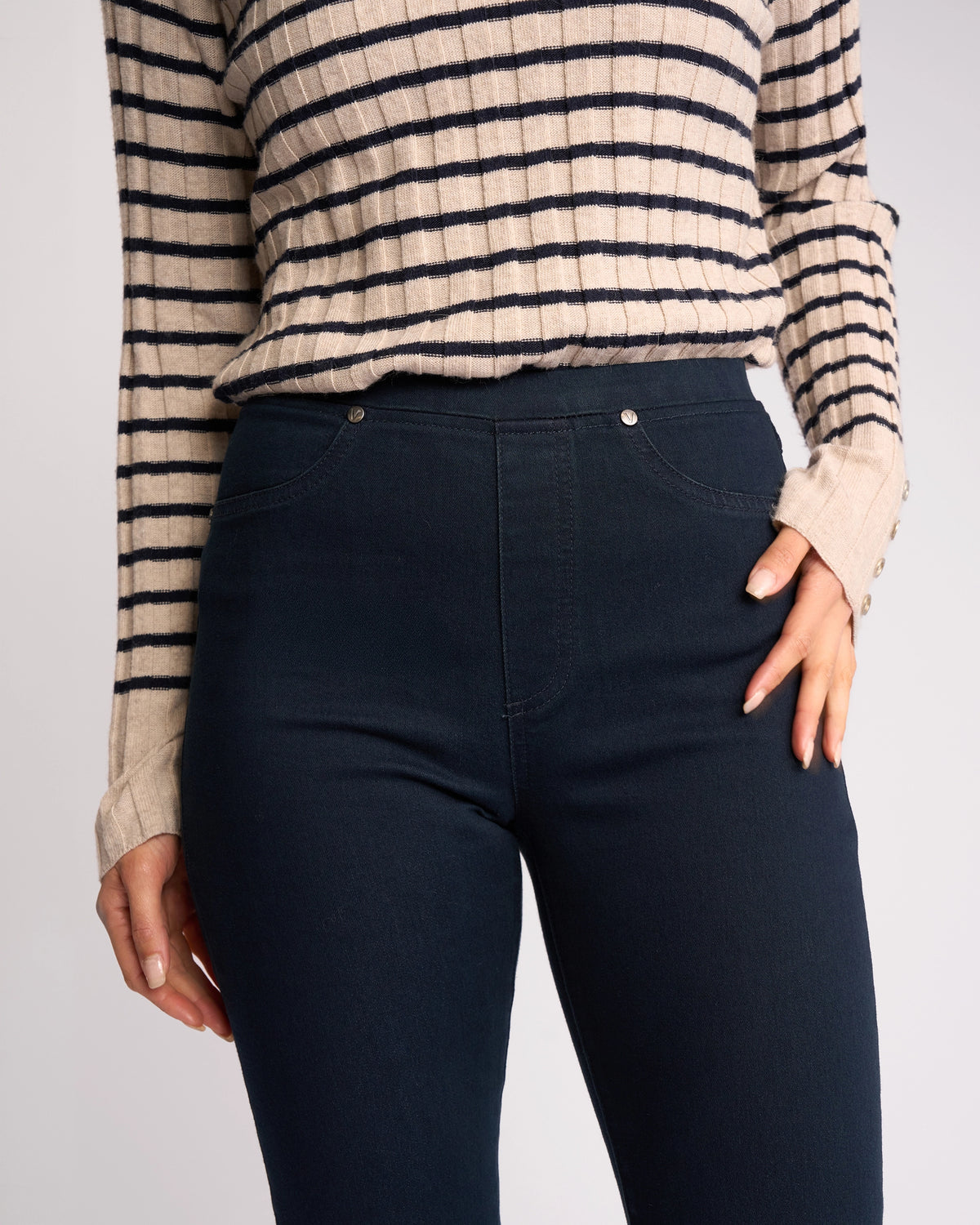 A person wears Vassalli's Bay Pull On - Indigo Denim jeans with a beige and black striped long-sleeve sweater, one hand on their hip. The image is cropped at the shoulders and upper thighs.