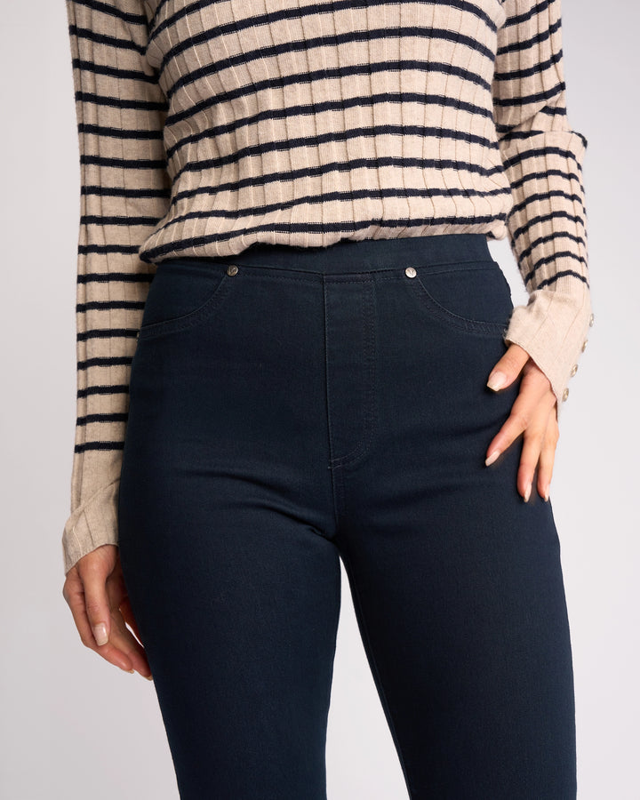 A person wears Vassalli's Bay Pull On - Indigo Denim jeans with a beige and black striped long-sleeve sweater, one hand on their hip. The image is cropped at the shoulders and upper thighs.