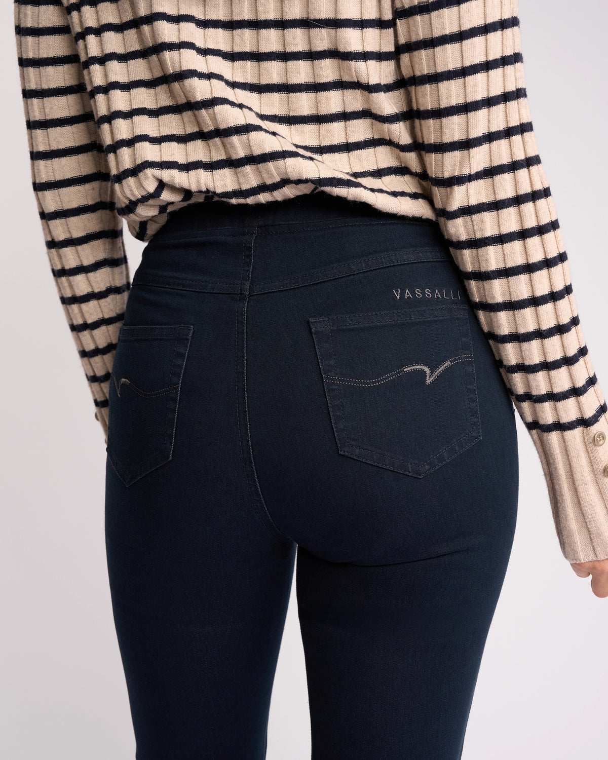 A person shown from the back wears a beige and black striped sweater and Vassalli Bay Pull On - Indigo Denim jeans. The slim-leg jeans feature stretch denim, two back pockets, and "VASSALLI" embroidered above the right pocket.