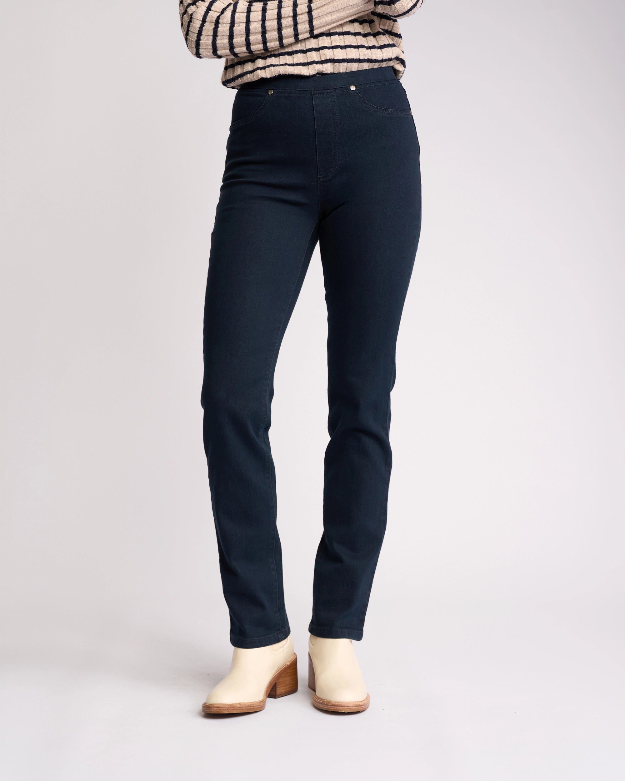 A person stands against a plain light gray background wearing Vassalli’s Bay Pull On - Indigo Denim jeans, a long-sleeved, light-striped shirt, and cream heeled ankle boots. Their arms are crossed and their head is not visible.