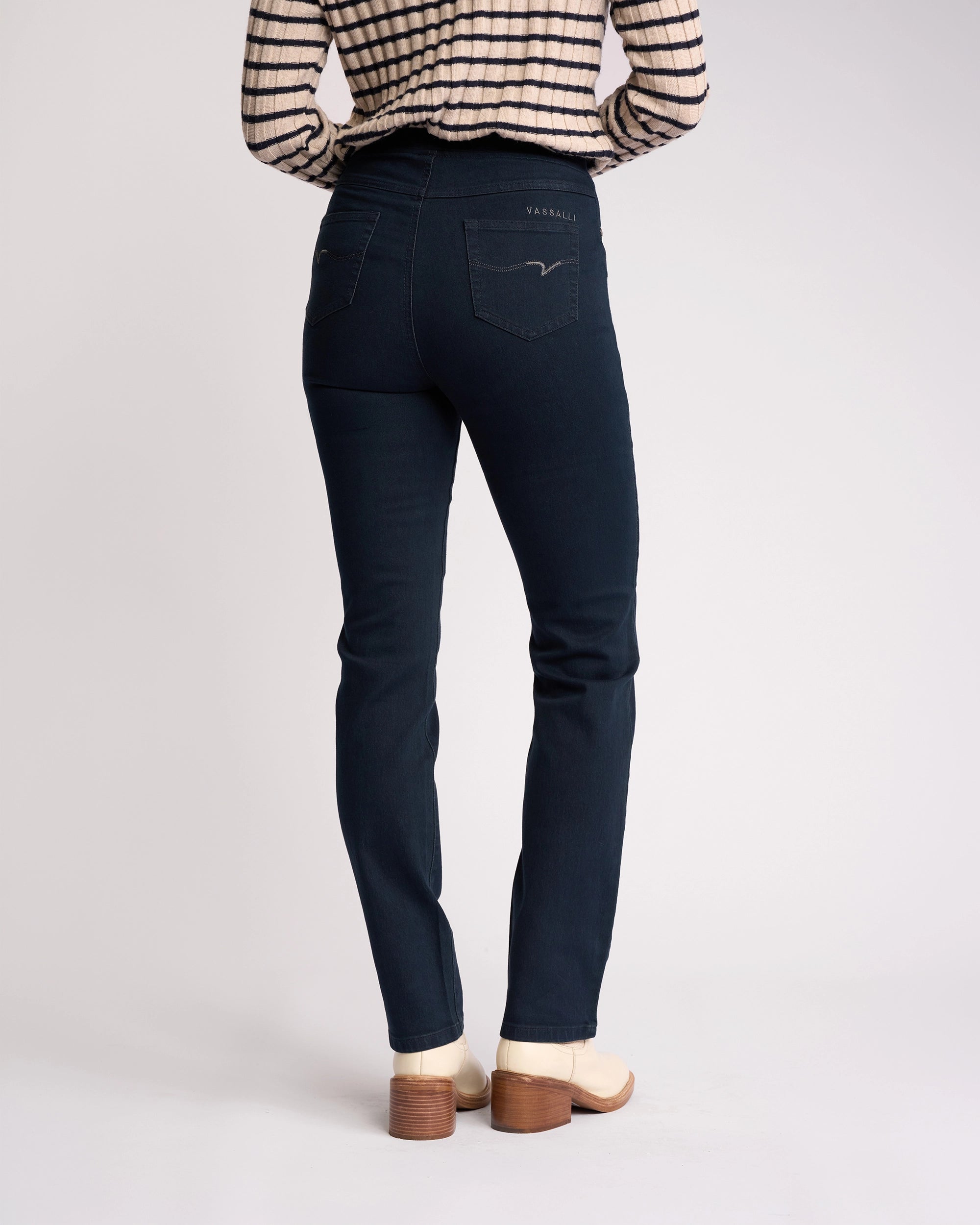 A person stands facing away from the camera against a plain light background, wearing a striped long-sleeve shirt and Vassalli Bay Pull On - Indigo Denim jeans, paired with light-colored heeled shoes.