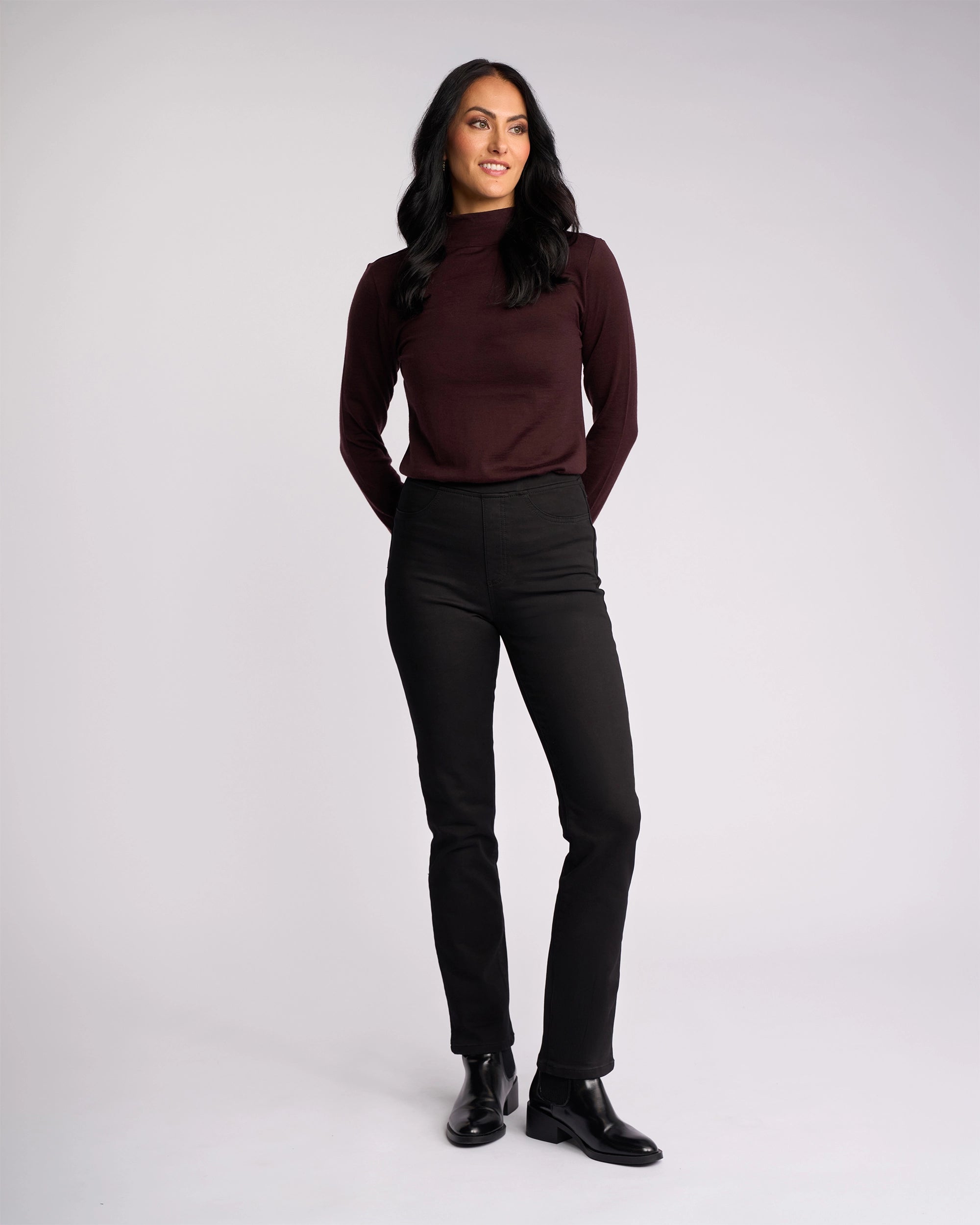 A woman with long dark hair stands against a plain background, smiling and looking to the side. She wears the Vassalli Bay Knit Pull On - Black pants, featuring a high-rise waist and slim-leg silhouette in comfortable stretch fabric.