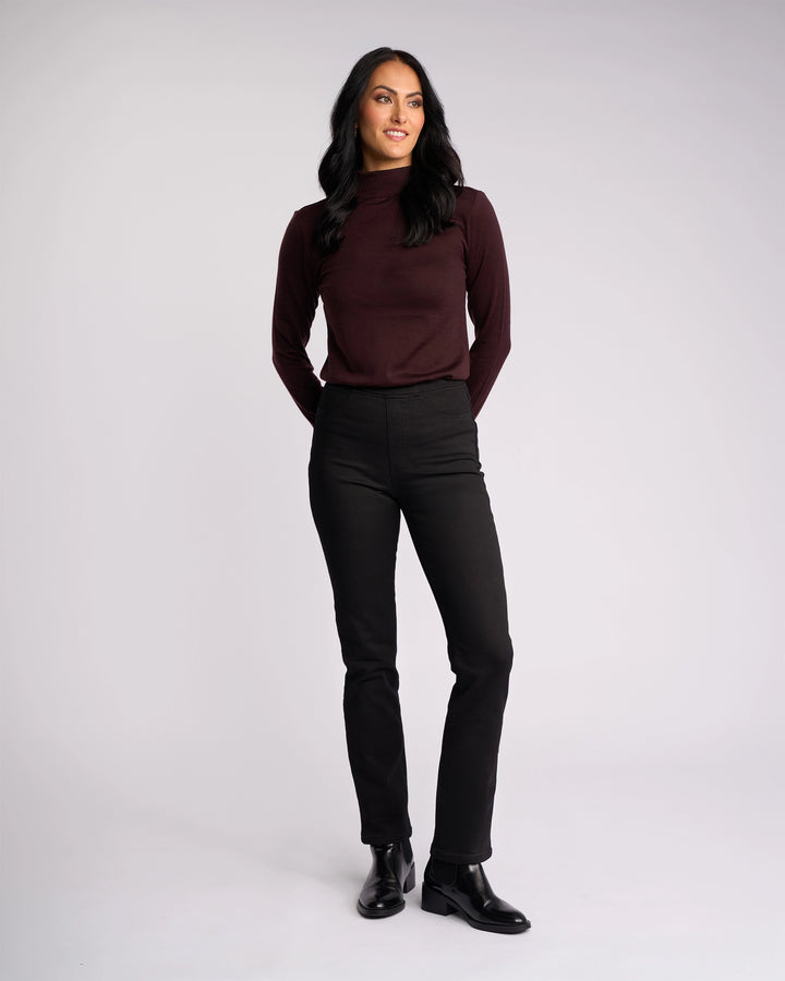 A woman with long dark hair stands against a plain background, smiling and looking to the side. She wears the Vassalli Bay Knit Pull On - Black pants, featuring a high-rise waist and slim-leg silhouette in comfortable stretch fabric.