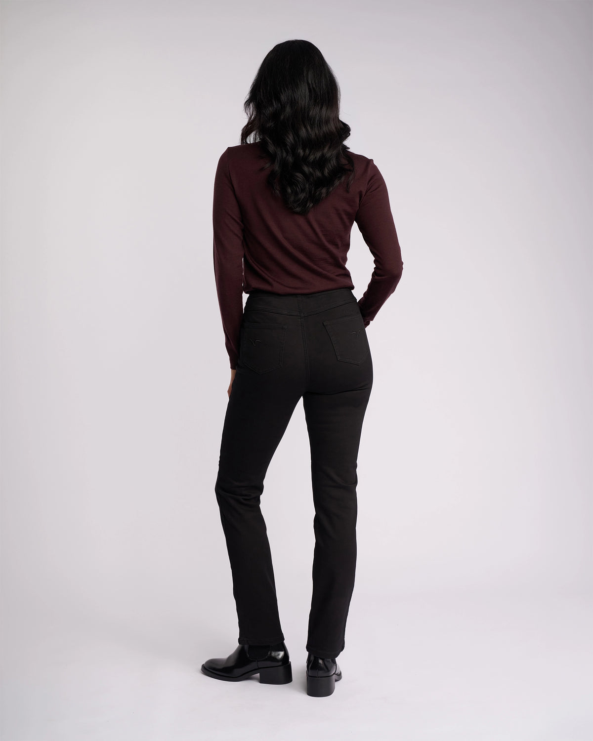 A person with long, wavy dark hair faces away in a maroon top, Vassalli Bay Knit Pull On - Black high-rise slim-leg pants, and black shoes, set against a plain white background.