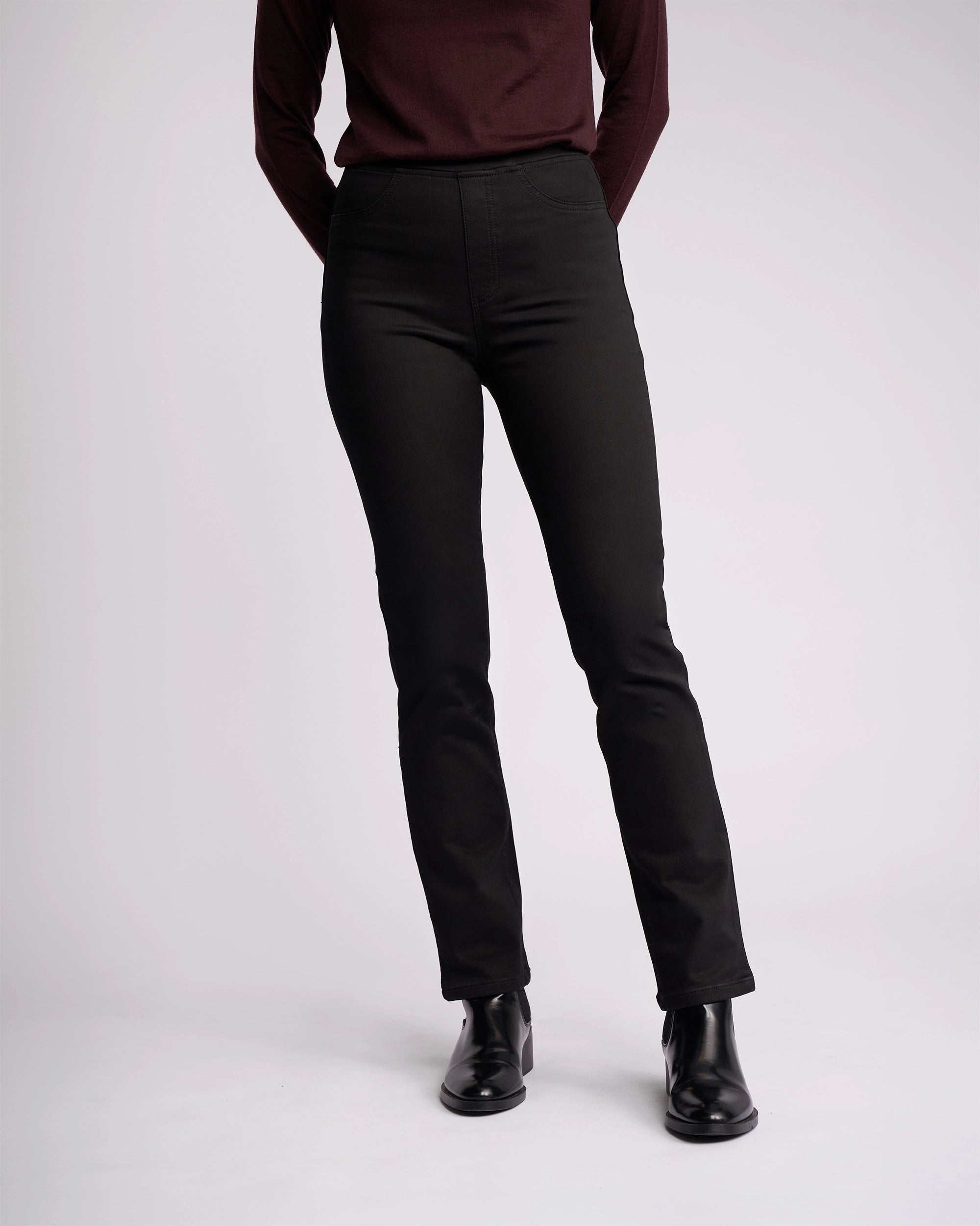 A person wears the Vassalli Bay Knit Pull On - Black pants with a long-sleeve burgundy top and black ankle boots, standing against a light background. The upper part of their face is not visible.