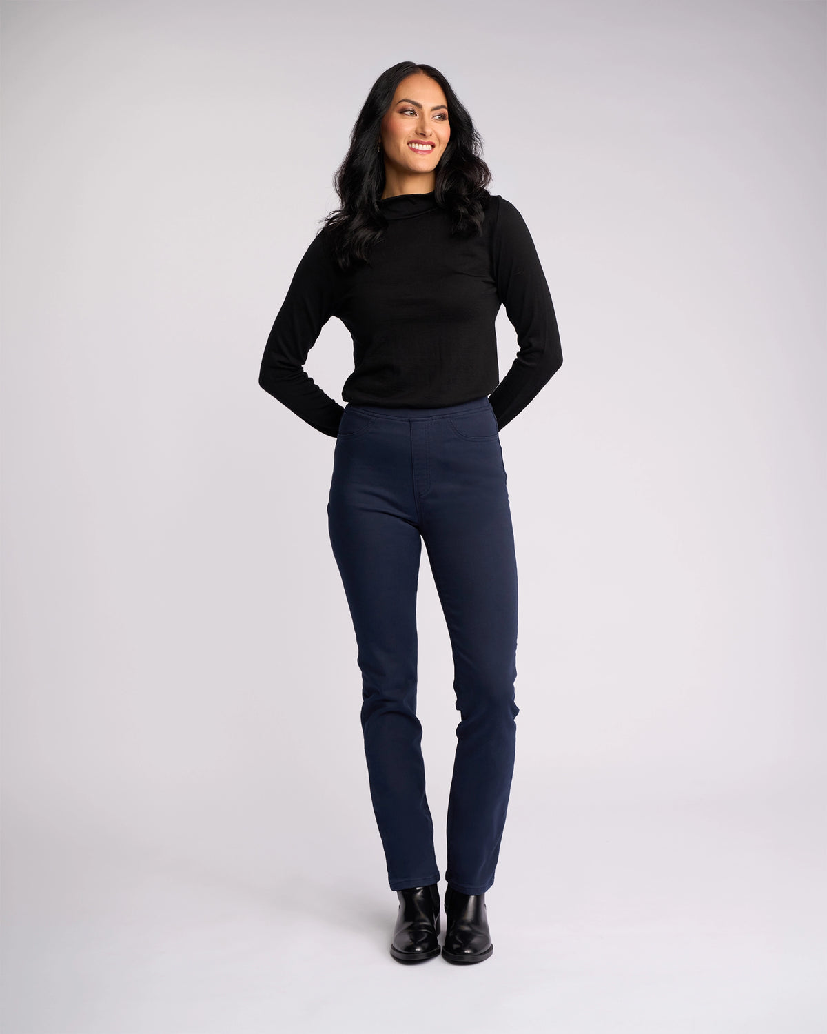 A woman with long dark hair smiles against a plain background, wearing the Vassalli Bay Knit Pull On - Carbon, a black long-sleeve top and dark blue high-rise slim-leg pants in stretch fabric, with her hands behind her back.
