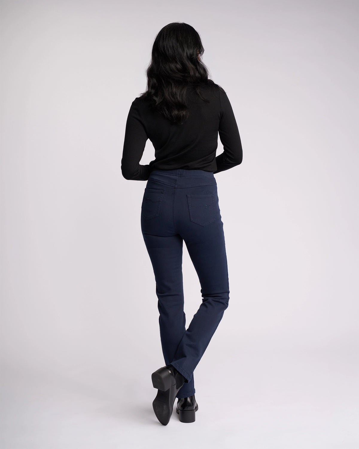 A person with long dark hair models the Vassalli Bay Knit Pull On - Carbon, a black stretch high-rise slim-leg pant, standing with one leg crossed behind the other against a plain light background.