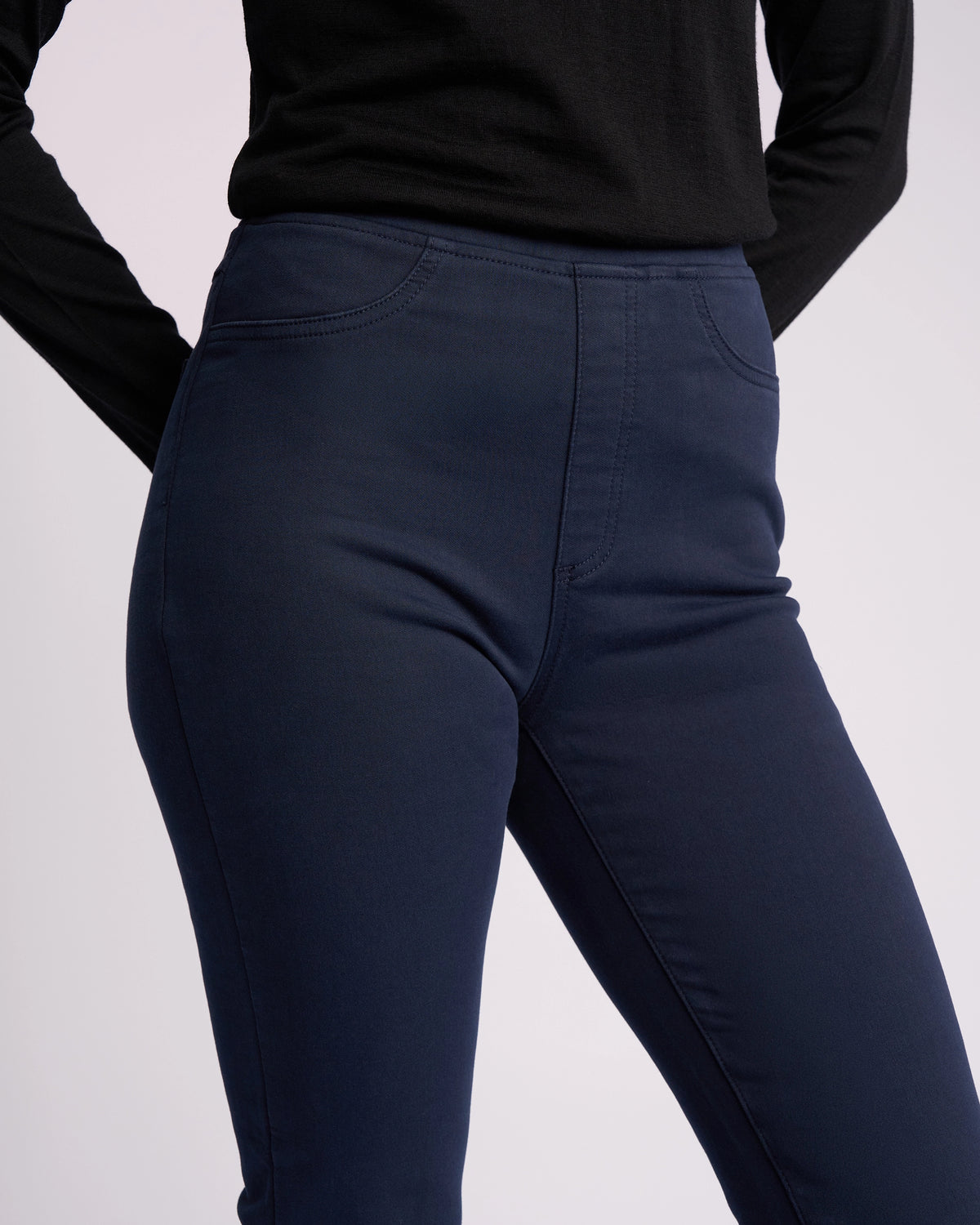 A person wears the Vassalli Bay Knit Pull On - Carbon, a black long-sleeve top and high-rise, dark blue slim-leg stretch pants, shown from shoulders to thighs with one arm bent and the other at their side.