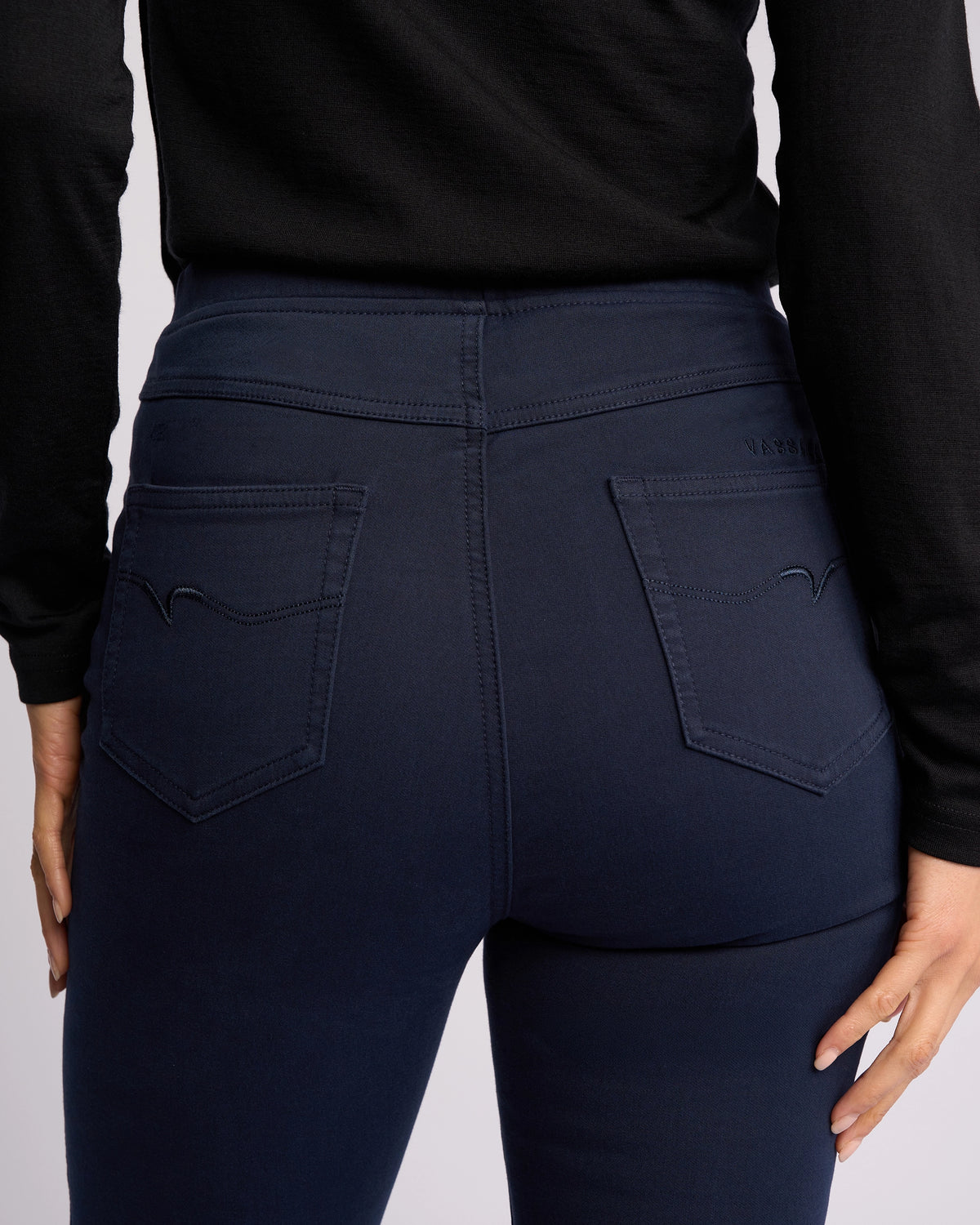 A person is shown from the back wearing Vassalli's Bay Knit Pull On - Carbon, a high-rise, dark blue slim-leg pant with two stitched back pockets. They pair it with a black long-sleeve top and have their hands at their sides.