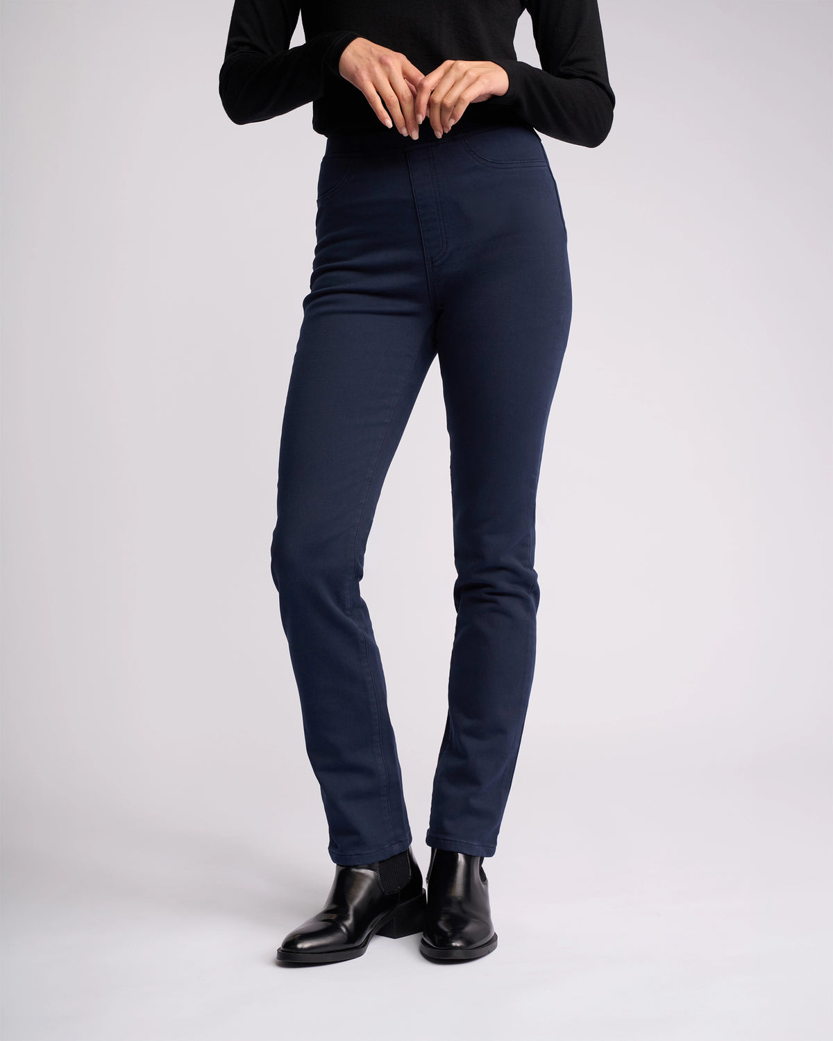 A person stands against a plain light background wearing the Vassalli Bay Knit Pull On - Carbon, a black long-sleeve top, and black ankle boots. The upper part of their face is not visible.
