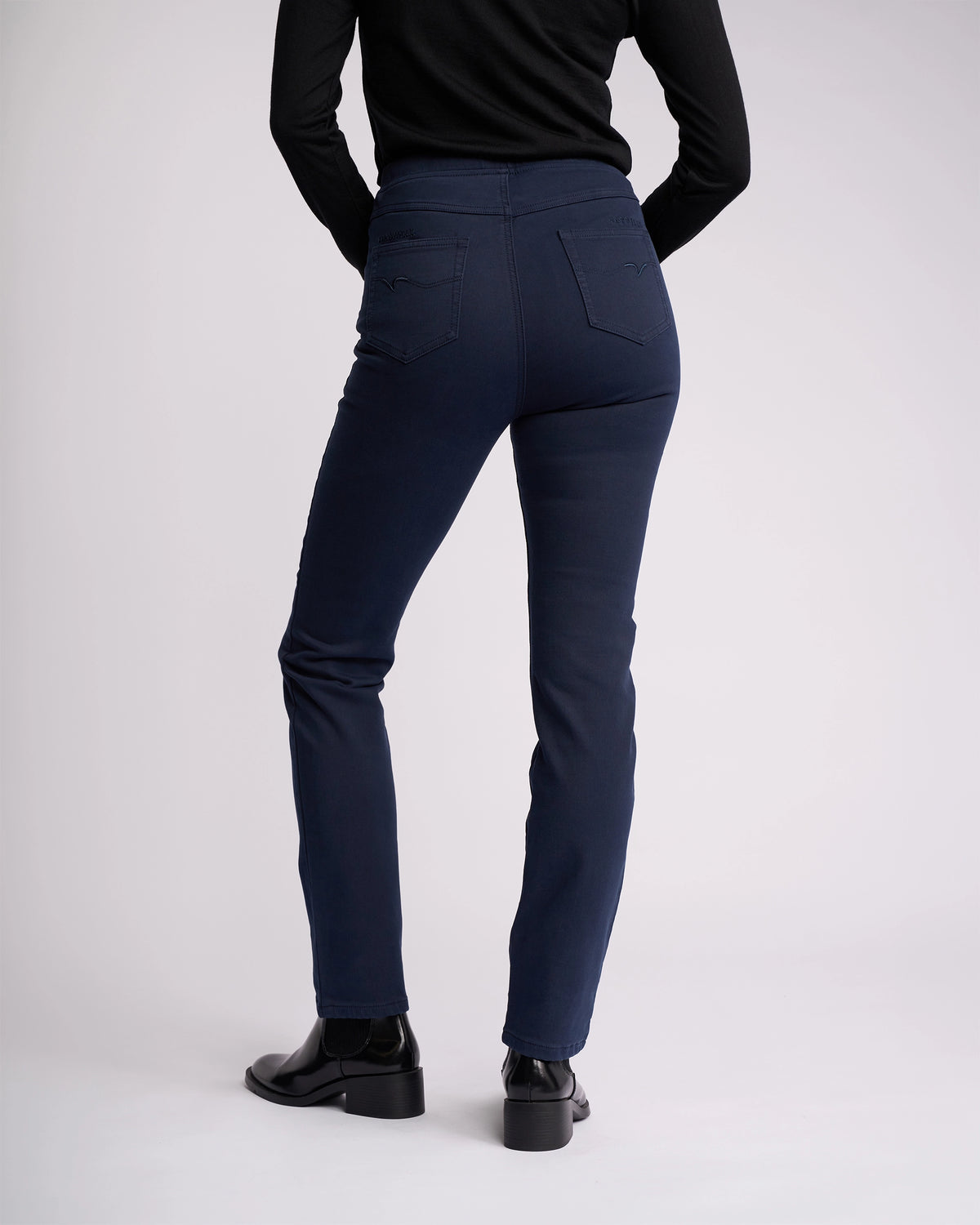 A person in the Vassalli Bay Knit Pull On - Carbon stands with their back to the camera, wearing a black long-sleeve top and black ankle boots against a plain light background.