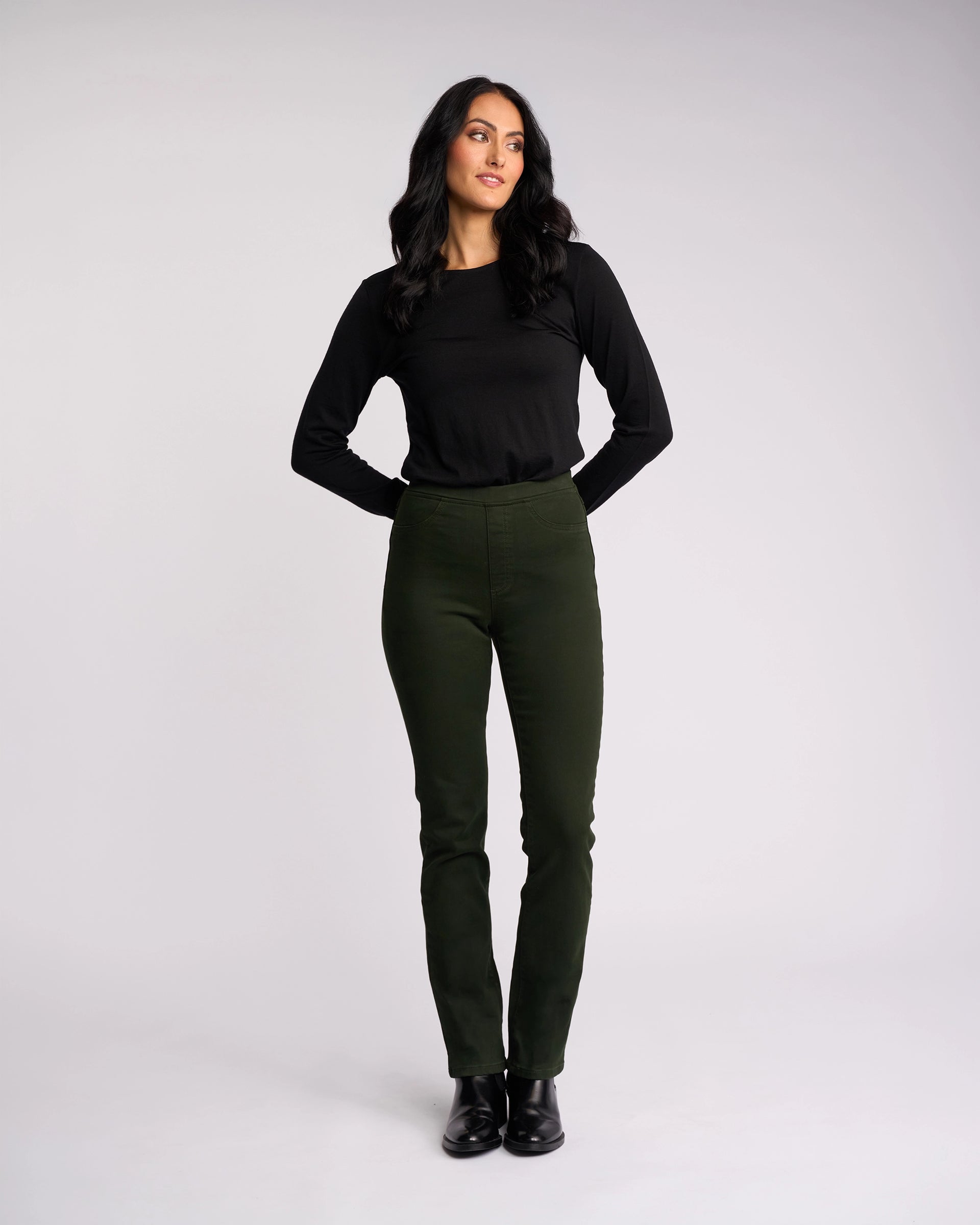 A woman with long dark hair stands against a plain backdrop, wearing the Vassalli Bay Knit Pull On pants in forest green and a black long-sleeve top. She looks slightly to the side with her hands behind her back.