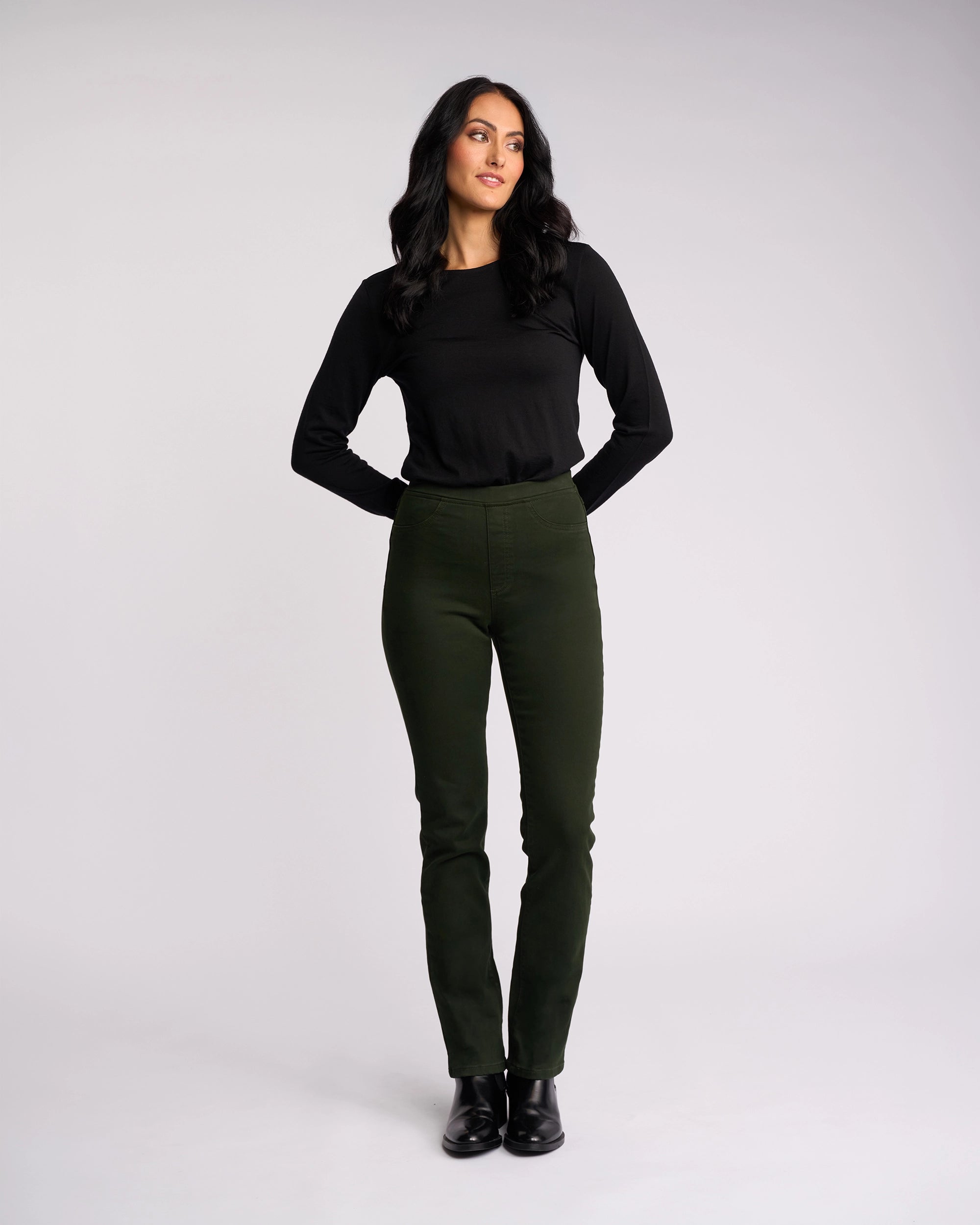 A woman with long dark hair stands against a plain backdrop, wearing the Vassalli Bay Knit Pull On pants in forest green and a black long-sleeve top. She looks slightly to the side with her hands behind her back.