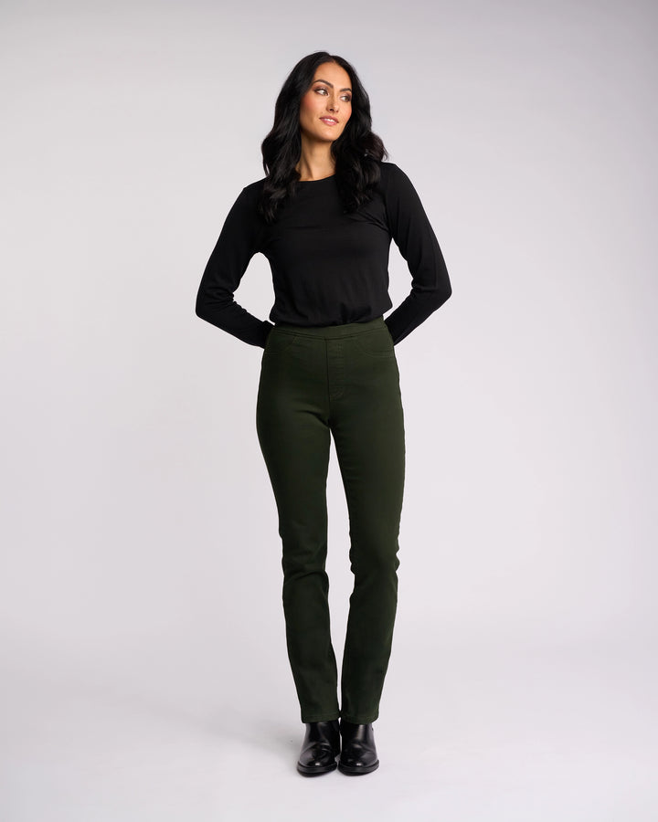 A woman with long dark hair stands against a plain backdrop, wearing the Vassalli Bay Knit Pull On pants in forest green and a black long-sleeve top. She looks slightly to the side with her hands behind her back.