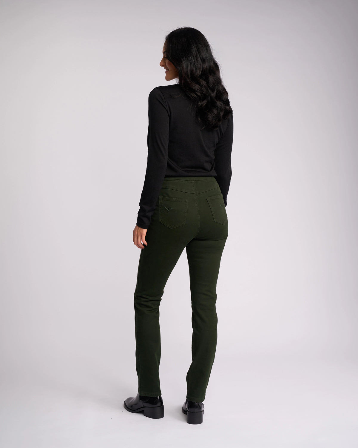 A woman with long dark hair, wearing the Vassalli Bay Knit Pull On in Forest, stands facing away from the camera against a plain light background.