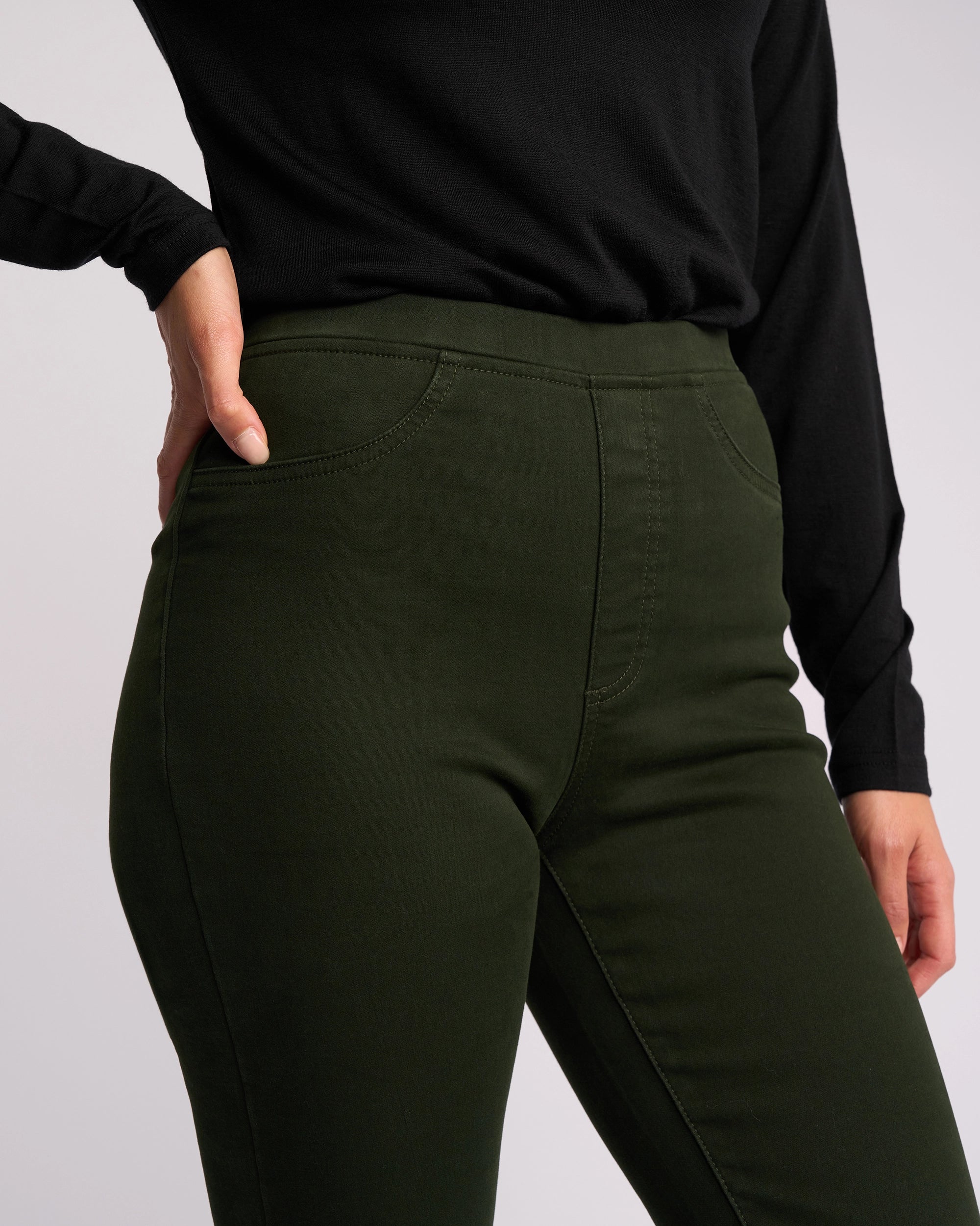 A person wears the Vassalli Bay Knit Pull On - Forest slim-leg pants with a black long-sleeve top, standing with one hand on their hip against a plain background.