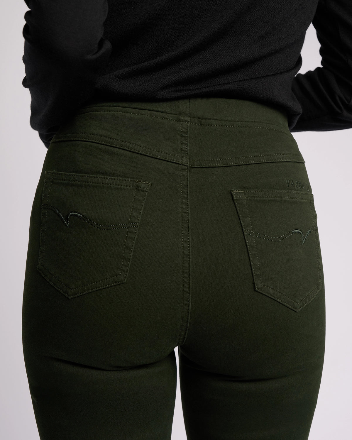 A person is shown from the waist down, back facing the camera, wearing Vassalli's Bay Knit Pull On - Forest. The dark green, slim-leg pants feature two back pockets with subtle stitching detail, paired with a black top.