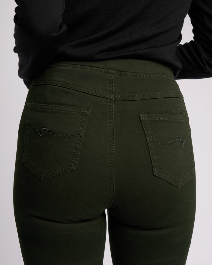 A person is shown from the waist down, back facing the camera, wearing Vassalli's Bay Knit Pull On - Forest. The dark green, slim-leg pants feature two back pockets with subtle stitching detail, paired with a black top.