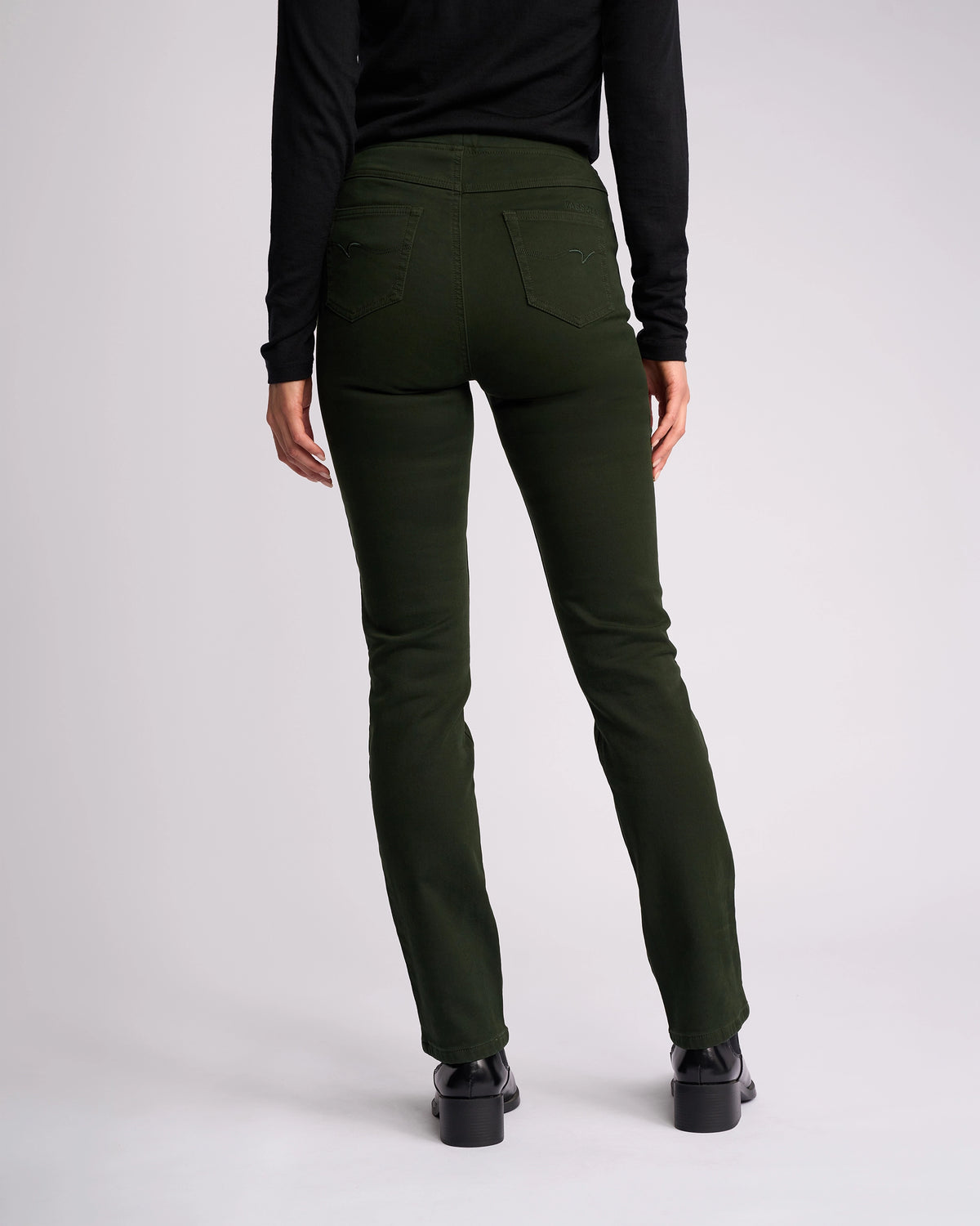 A person in Vassalli's Bay Knit Pull On - Forest, a dark green high-rise slim-leg pant, stands with their back to the camera wearing a black long-sleeve top against a plain light background.