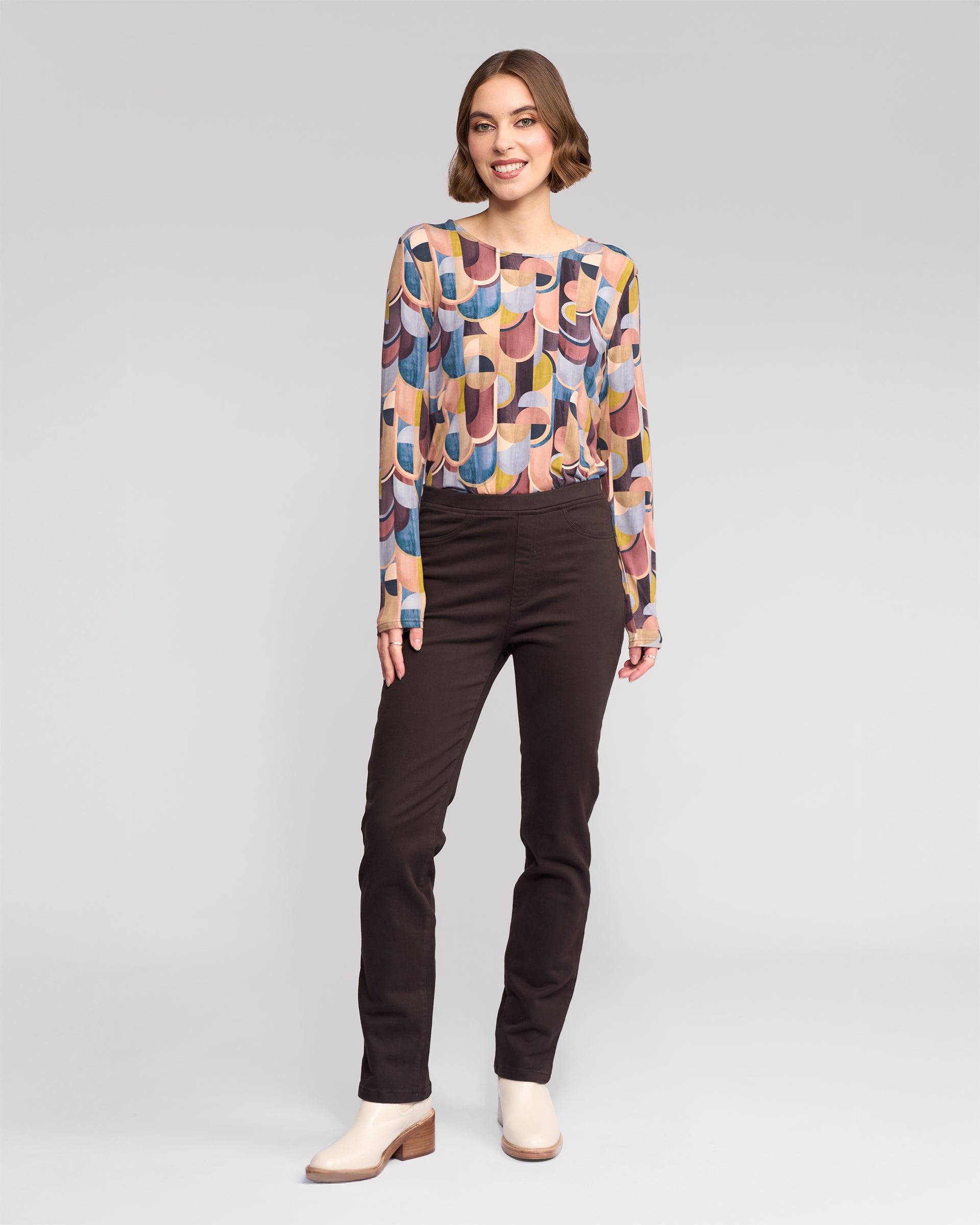 A woman smiles in a studio, pairing the Vassalli Bay Knit Pull On - Chocolate pants with a colorful geometric top and cream ankle boots, all set against a plain gray background.