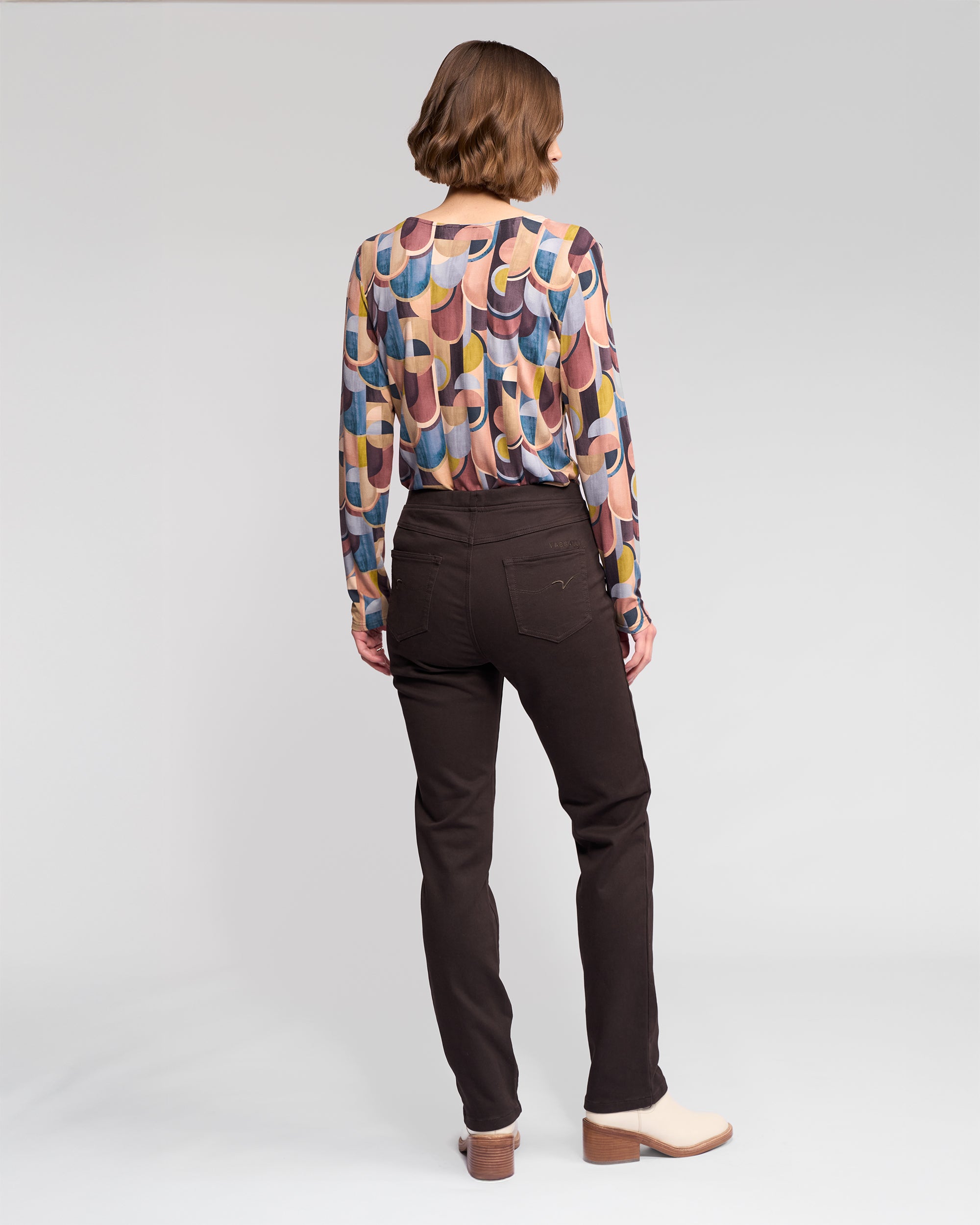 A woman stands facing away in Vassalli’s Bay Knit Pull On - Chocolate slim pant, a colorful patterned long-sleeve top, and beige heeled shoes against a plain light gray background.